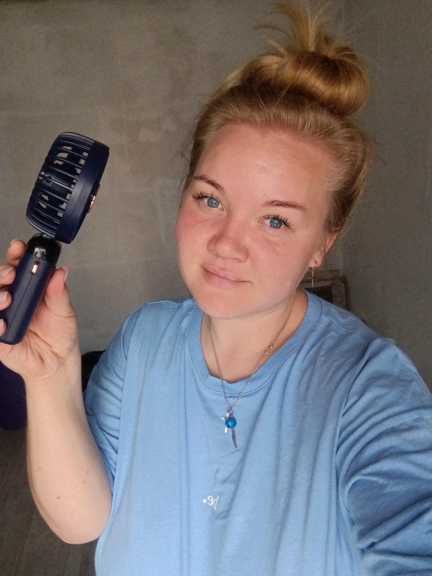 In a dimly lit indoor setting, a woman with blonde hair tied in a bun and blue eyes holds a dark blue portable fan in her right hand. She is wearing a light blue t-shirt and a silver necklace with a blue pendant. Her left arm is visible, and she has a slight smile on her face. The background appears to be a wall with a textured surface. The scene is captured from the chest up.
