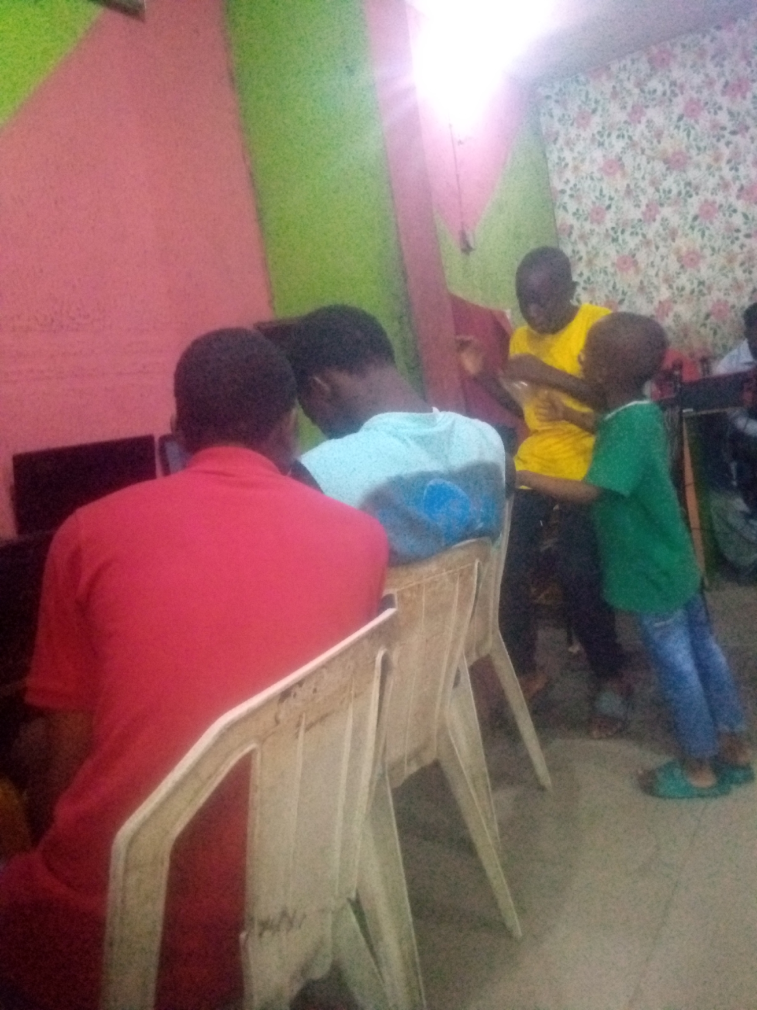 In a room in Shangisha, Nigeria, three young males and one adult male are engaged in activities. One young male, wearing a yellow t-shirt, stands and gestures. Another young male, in a green t-shirt and blue jeans, stands facing him. In the foreground, an adult male in a red shirt is seated at a computer, with his back to the camera. To his left, another male in a light blue t-shirt is also seated, facing the computer. The room has pink and green walls and floral wallpaper.