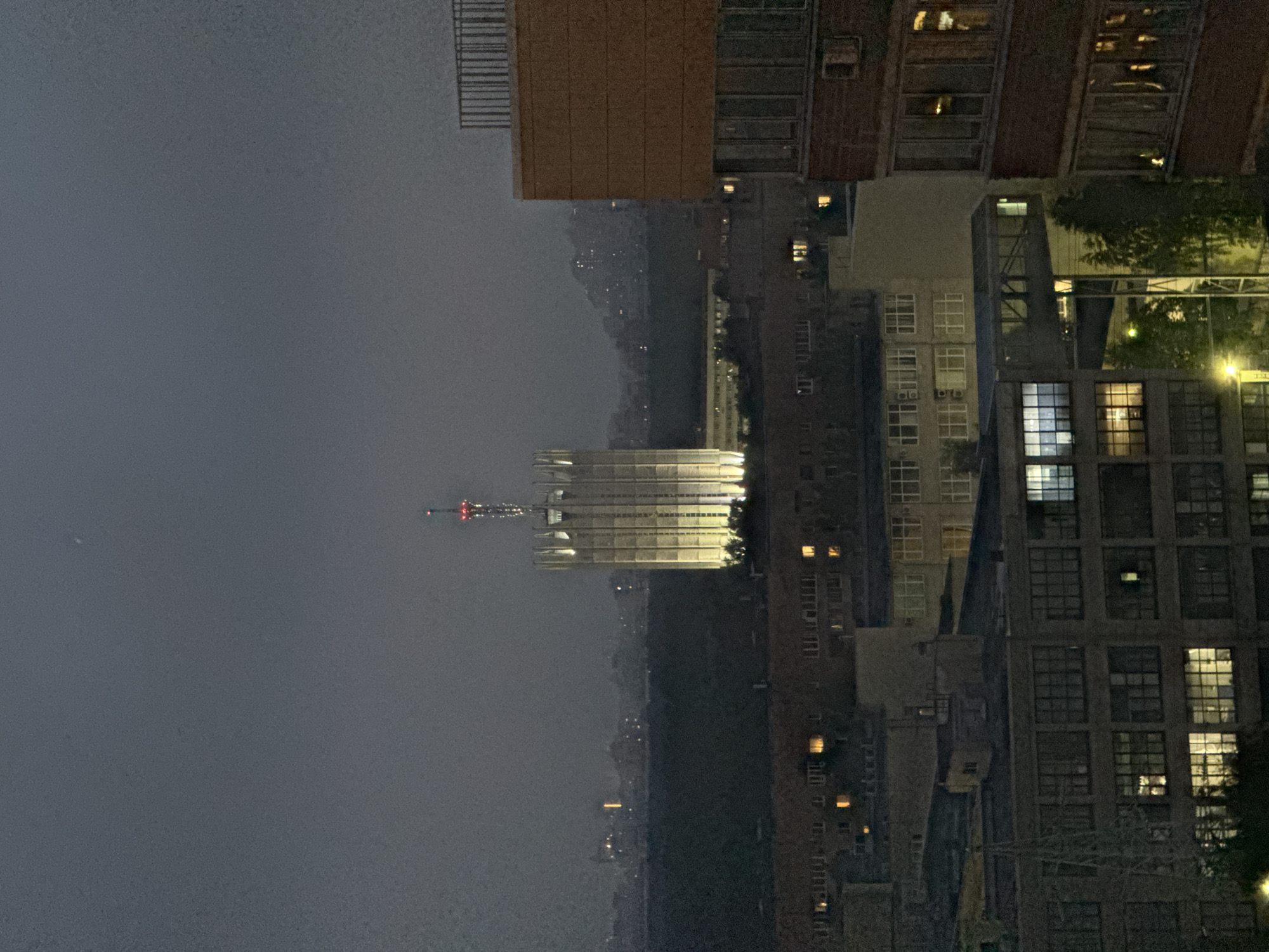 The image captures an urban landscape at dusk or night. In the foreground, the side of a multi-story apartment building is visible, with illuminated windows suggesting occupancy. To the left of this building, another building with large windows and industrial-style architecture is seen. A metal tower-like structure is partially visible in front of this building.
Dominating the mid-ground and background is a tall, illuminated tower with a distinct architectural style featuring vertical fluting and a spired top adorned with red lights. This tower is surrounded by a dark expanse of trees and other urban structures, some with faint lights. In the far distance, a hazy silhouette of a city or town with scattered lights is visible against the dim sky, which has a single bright speck that could be a star or a distant light. The overall atmosphere is muted and dark, with artificial lighting from the buildings and the tower being the primary sources of illumination.