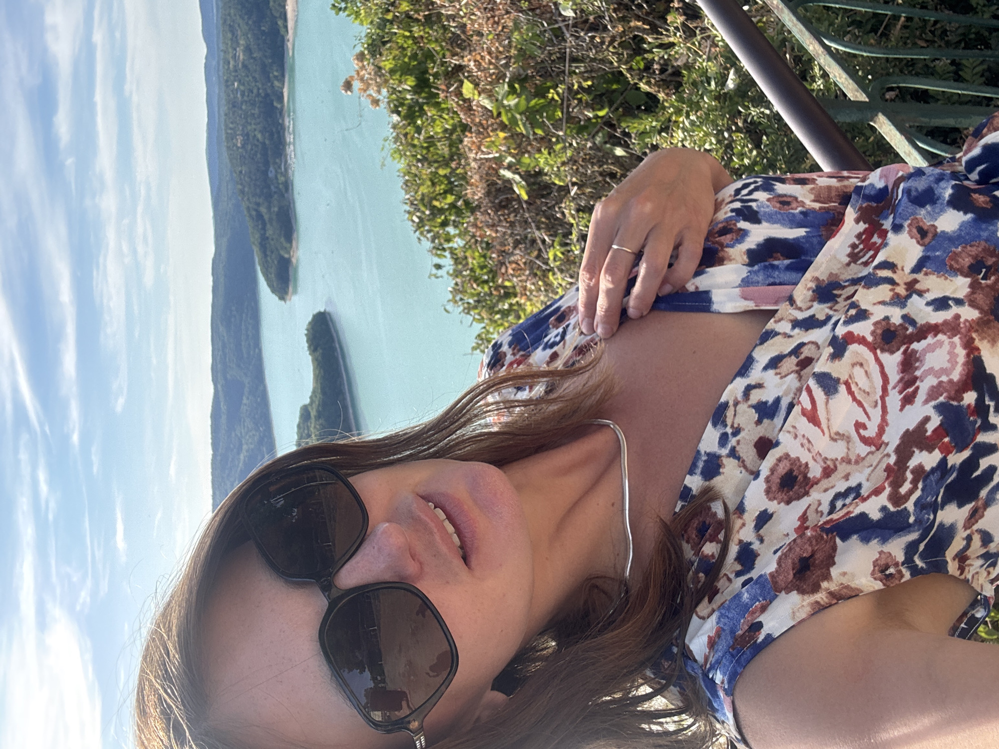 In Moirans-en-Montagne, France, a woman wearing large, dark sunglasses and a floral dress poses for a selfie against a backdrop of a bright blue lake surrounded by lush, green hills under a partly cloudy sky. She has brown hair and is wearing a silver necklace. Her right hand is near her chest, and she has a ring on her right ring finger. The woman is standing next to a railing with green metalwork, suggesting she is on a balcony or viewpoint. The scene is set outdoors, likely on a sunny day.