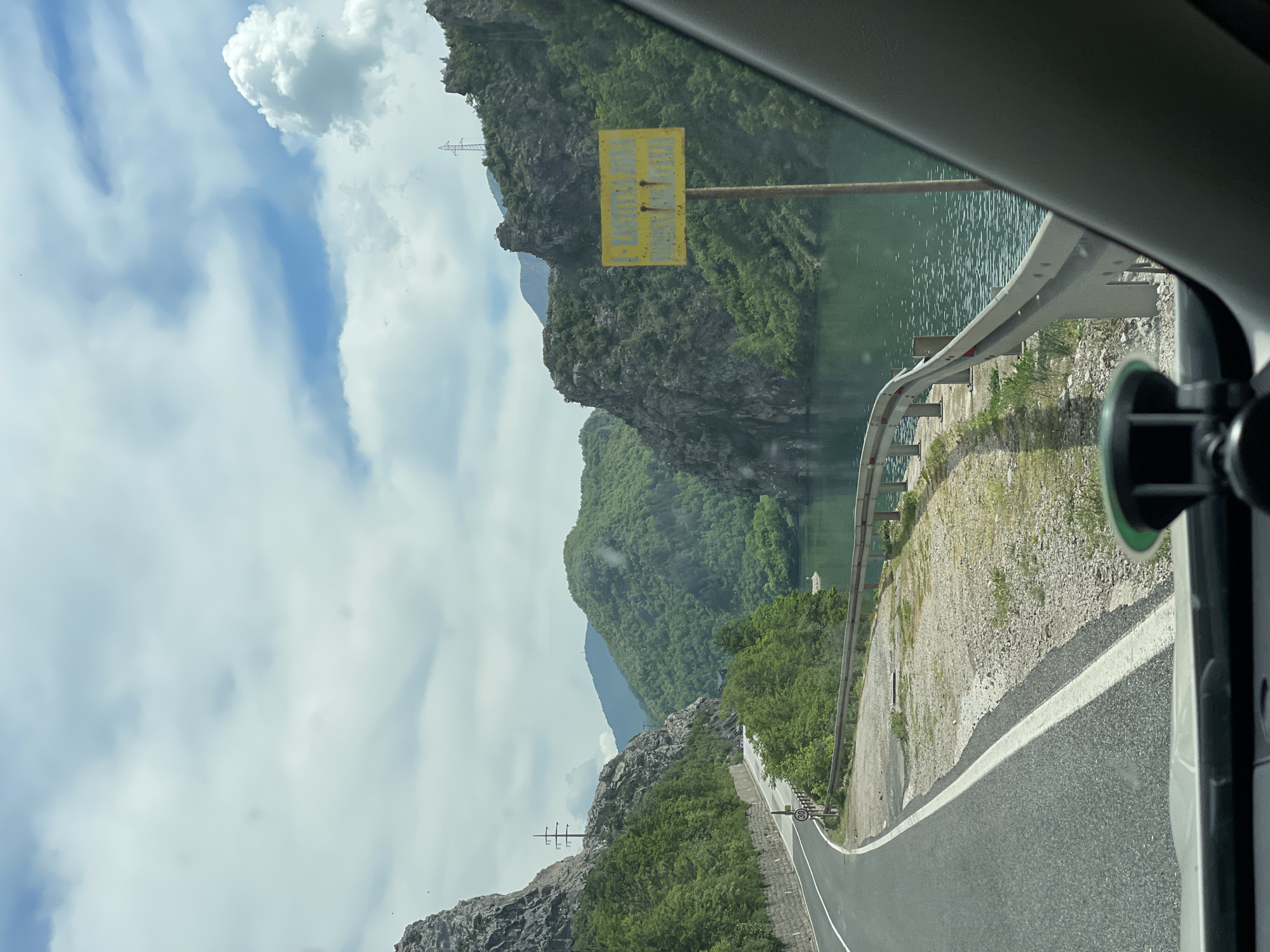 The image captures a view from inside a vehicle, looking out onto a winding road in Glogošnica, Bosnia and Herzegovina. The road is bordered by a safety guardrail on the right and is adjacent to a body of water, likely a lake or river. Lush greenery and rocky cliffs characterize the surrounding landscape. The sky is filled with a mix of blue patches and fluffy white clouds. A yellow sign is visible on the left side of the road, affixed to a post. A suction-cup-mounted device is attached to the inside of the vehicle window. The scene suggests a scenic drive through a mountainous and forested region.