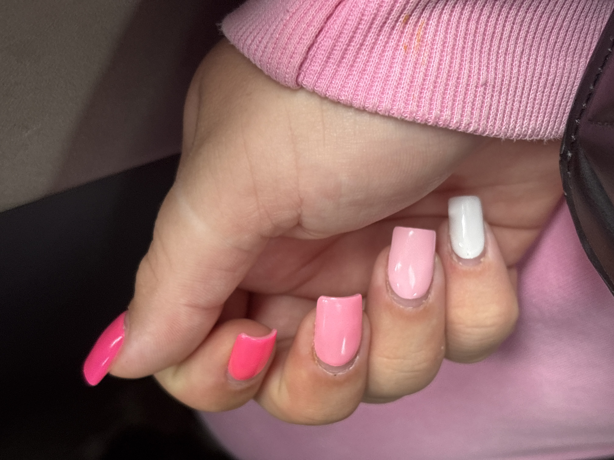A close-up shot shows a person's left hand with bright pink and light pink square-shaped acrylic nails, as well as a white nail. The hand is resting against a pink fabric garment. The fingers are slightly curled. The background is dark. The image appears to be taken indoors, possibly in a car or on a couch. No specific location in Mahilyow, Belarus, can be identified from this image.