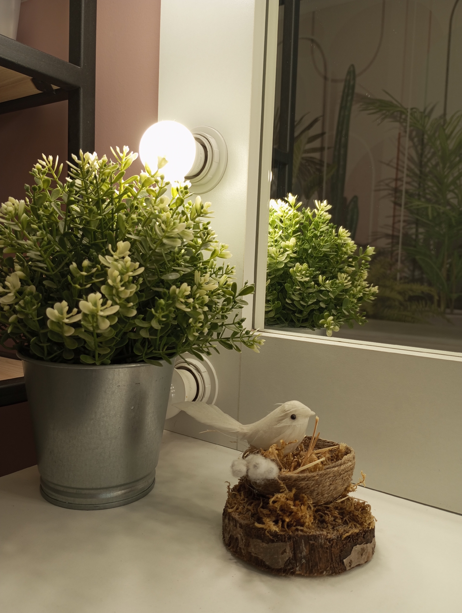 The image depicts a decorative indoor setting. A silver pot filled with a faux green plant sits to the left. Beside it, there is a decorative bird nest on a small piece of tree bark with a white, decorative bird on top, complete with some cotton balls beside it. The background features a mirror reflecting the plant and what looks like a cactus. The area is illuminated by a light bulb, which is attached to a white wall. The setting appears to be on a white tabletop. The location in Izhevsk, Russia is not discernible from this image.