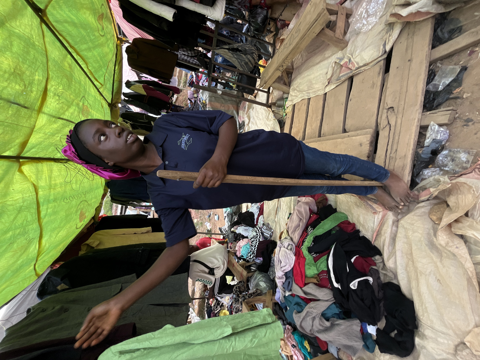 A young person, likely a vendor, lies on their back under a bright green tarp, holding a wooden pole. They are wearing a dark blue polo shirt with a visible logo and blue jeans. Their head is tilted up, looking towards the sky. The scene appears to be a market stall in Nyanya, Nigeria. Piles of colorful clothing are scattered around, some neatly stacked and others in disarray. Wooden planks and makeshift shelving are visible, suggesting a rustic and perhaps temporary setup. The overall impression is one of a busy, possibly dusty, open-air market. The lighting suggests it could be daytime, and the presence of outdoor elements hints at fair weather. The young person's expression is difficult to discern, but their posture suggests a moment of rest or perhaps engagement with their merchandise.