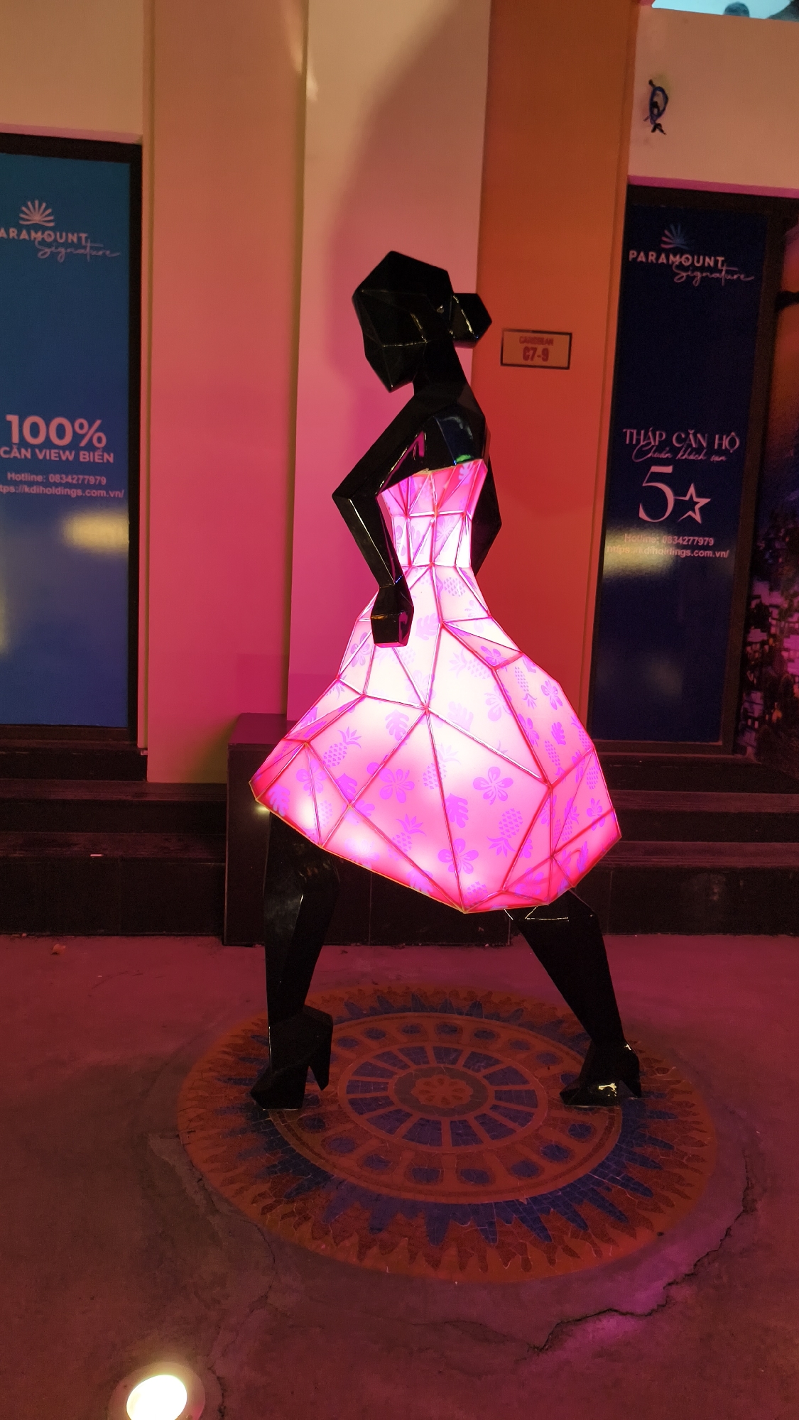 The image shows a life-sized, stylized black sculpture of a woman wearing a geometric, illuminated pink dress with floral and pineapple patterns. The sculpture stands on a circular mosaic tile floor. The background includes a pink wall and two dark blue signs with white text. One sign, on the left, has a logo and text that reads "PARAMOUNT Signature" and "100% CÀN VIEW BIỂN," followed by a hotline number and website. The other sign, on the right, also features "PARAMOUNT Signature" and text that appears to be in Vietnamese, including "THÁP CĂN HỘ 5*." A small sign with "C7-9" is visible on the wall between the two larger signs. The lighting in the scene is predominantly pink and red, with a spotlight visible on the ground in the lower left corner. The overall environment suggests an outdoor or semi-outdoor public space, possibly in a commercial or residential complex in Bac Nha Trang Ward, Vietnam.