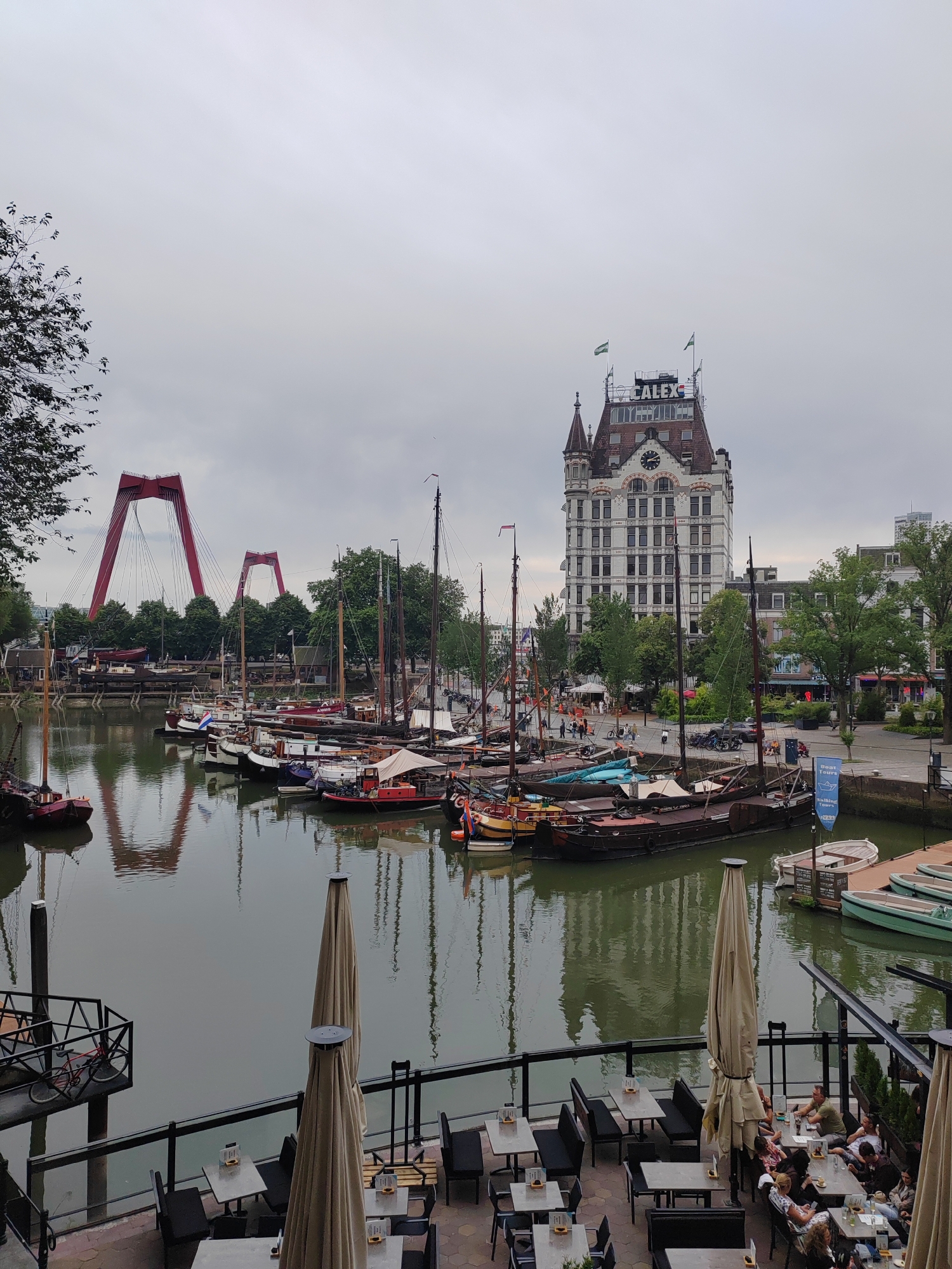 This is an outdoor shot of a harbor in Rotterdam, Netherlands, featuring a variety of boats docked on calm water. In the background, a prominent white building with "CALEX" signage on top stands against a cloudy sky. Two red suspension bridges are visible in the distance, their distinctive shapes a landmark of the city. The foreground shows an outdoor seating area of a restaurant or cafe with tables and chairs, some of which are occupied by people. Two large, closed umbrellas are set up to provide shade. The scene appears to be during the daytime, with overcast weather. The overall mood is peaceful, with the reflections of the boats and buildings creating a sense of tranquility.