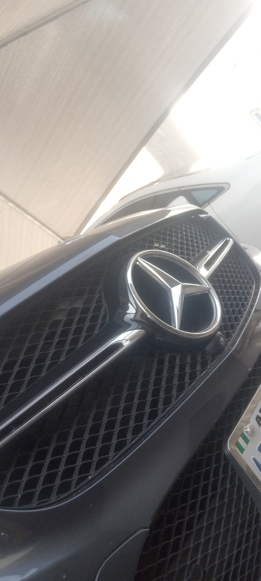 A close-up, angled view of the front of a dark-colored Mercedes-Benz. The Mercedes-Benz logo is prominently displayed on the grill. The vehicle is outdoors, possibly under a carport or awning, based on the roof structure visible in the background. A Nigerian license plate is partially visible. The scene appears to be in Narayi, Nigeria, based on the license plate.