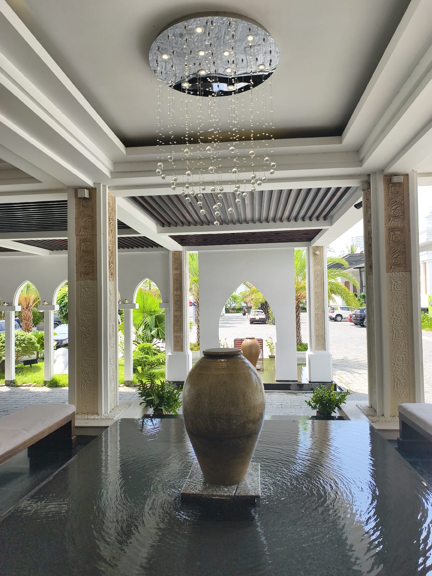 The image depicts the interior of a building in Bac Nha Trang Ward, Vietnam. A large ceramic vase sits in the center of a shallow pool of water, positioned beneath a round chandelier with hanging crystal-like beads. The building features white columns with carved designs, framing arched doorways that offer views of palm trees, other greenery, and a street with parked cars. Benches are visible on either side of the water feature. The ceiling has a coffered design with dark wood slats.