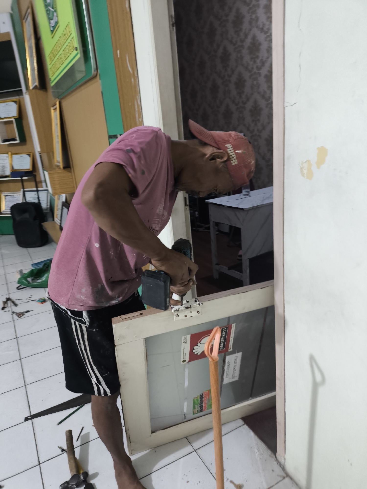 A man in a pink t-shirt and black shorts is using a power drill to attach a hinge to a window frame. He is wearing a red baseball cap. The scene appears to be indoors, possibly a room undergoing renovation or repair. In the background, there are framed documents on the wall and a piece of furniture. Tools such as a hammer and a saw are visible on the floor. The floor is tiled.