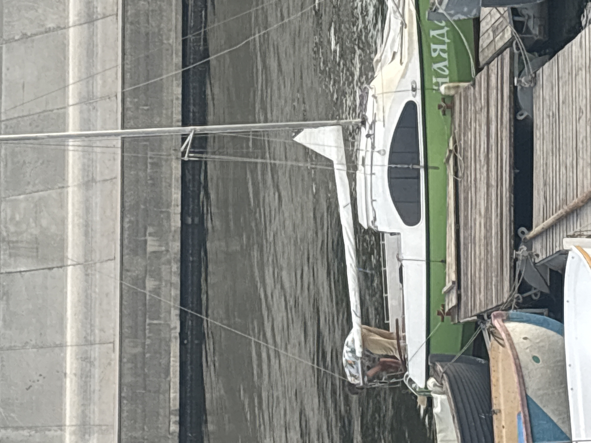 The image shows a small sailboat docked at a pier. A person wearing a patterned shirt and tan pants is leaning over the side of the boat, possibly working on something. The boat has a white hull with a green waterline and the word "НАДЯ" (Nadya) is visible on the side. Several other boats are also docked nearby. The background consists of a concrete wall and dark water. The scene appears to be in Astrakhan, Russia, given the context.