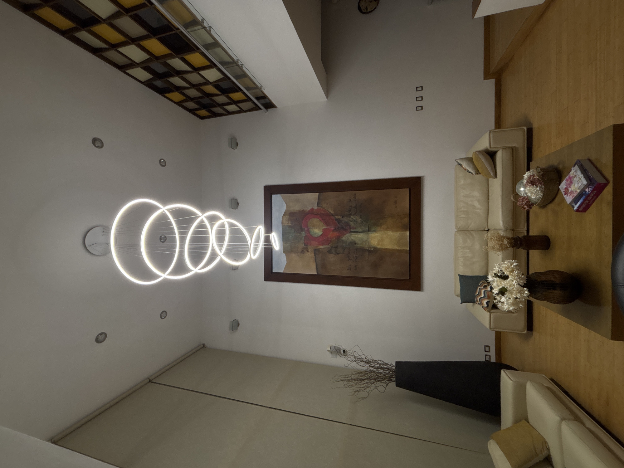 This media file captures a contemporary interior living space, likely a living room, in San Bernardino Tlaxcalancingo, Mexico. The room is brightly lit by a distinctive, modern chandelier composed of several glowing white circular rings suspended from the ceiling. Numerous recessed pot lights are also visible in the white ceiling, which features varying angles and levels.
The main wall displays a large, framed abstract painting with warm tones of red, orange, and brown, and what appears to be faint, cursive Spanish text incorporated into the artwork. Below the painting, a light-colored (possibly cream or beige) sofa with throw pillows is visible, suggesting a seating area. Further to the right, another similar sofa is partially seen.
In the foreground, a large, dark rectangular coffee table or console table holds decorative items, including a vase with white and dried flowers, and two books. One book has a floral cover, while the spine of the other clearly reads "INTERIOR DESIGN." A tall, slender black floor vase containing sparse, dried branches stands against the wall. The flooring appears to be light-colored wood.
The ceiling also features a unique multi-paneled section with a grid pattern of yellow, white, and black squares, possibly a decorative light fixture or skylight. The room is devoid of people, and no specific activity is taking place. The overall impression is one of a clean, well-designed, and modern residential or commercial interior, likely photographed in the evening or at night given the prominence of artificial lighting.