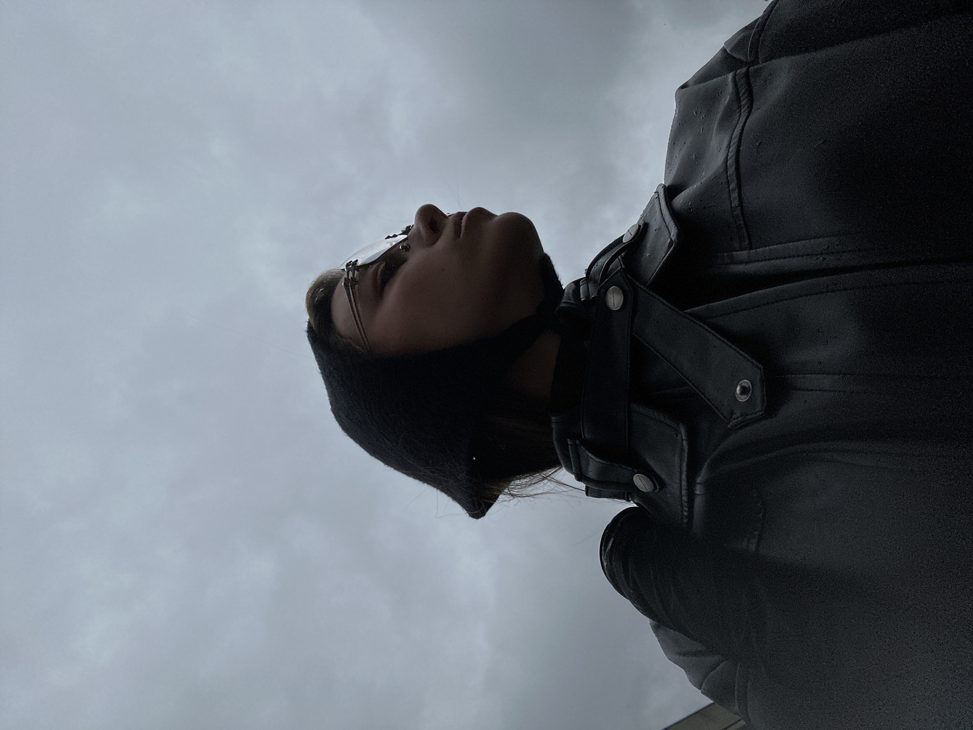 A low-angle, close-up, full-body shot captures a young person from the shoulders up, looking to the right against a cloudy sky. The subject, wearing a black knit cap and a black leather jacket, has on wire-rimmed glasses. The jacket appears to be wet, with visible water droplets. The background consists of a overcast, grey sky with subtle cloud formations. The image does not provide any discernible details about the location in Moscow, Russia.