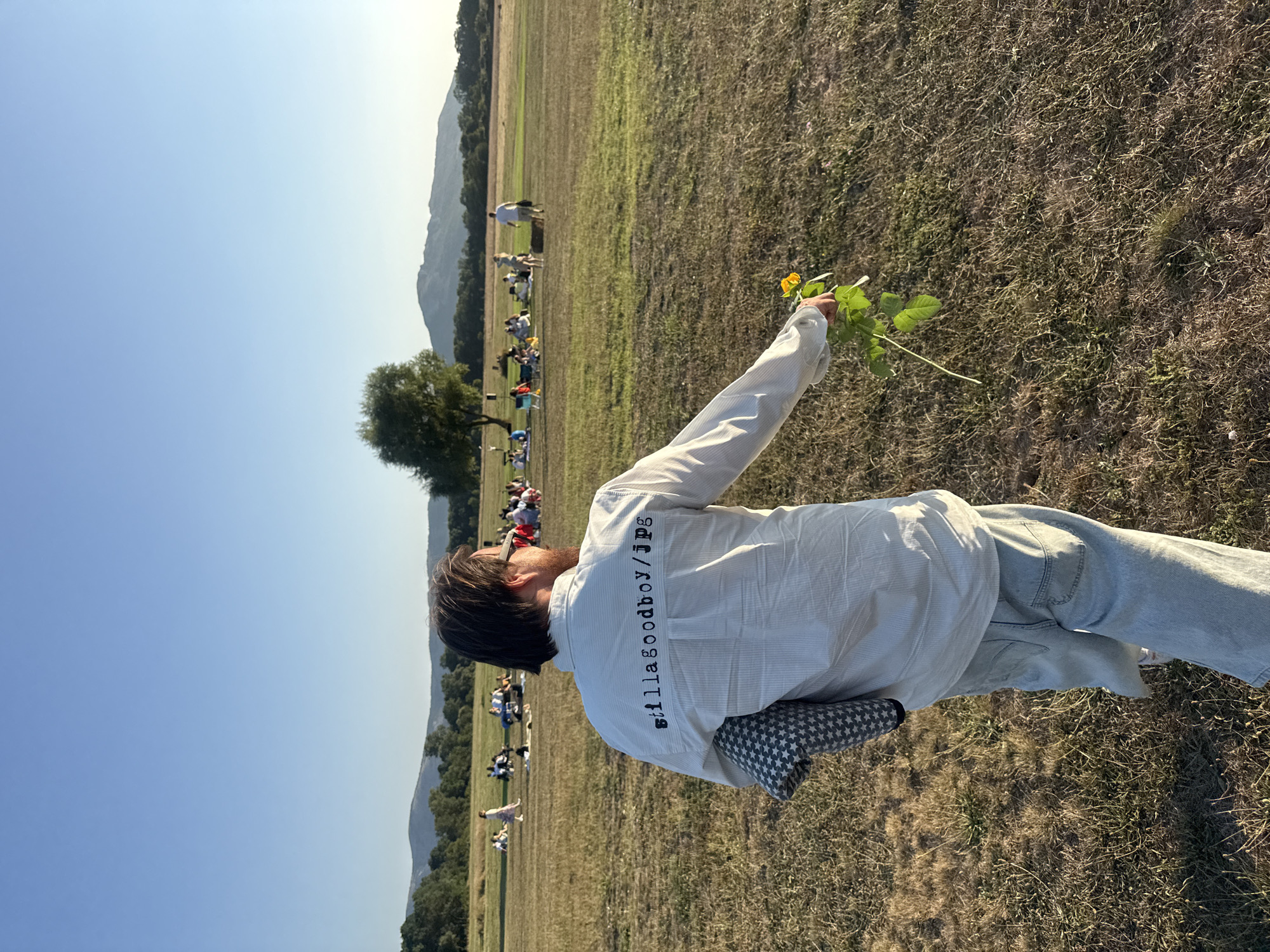 In a grassy field under a clear blue sky, a person stands with their back to the camera, holding a small yellow flower and a patterned blanket. The person is wearing a light-colored striped shirt with the text "stillagoodboy/jpg" printed on the back and light-wash denim jeans. In the background, a group of people are scattered across the field, some sitting on blankets and chairs, enjoying a gathering. A lone tree stands in the mid-ground, and rolling hills with trees and vegetation form the horizon. The scene appears to be outdoors in a rural setting, possibly on the outskirts of Орлиновский округ, Ukraine, during daylight hours.