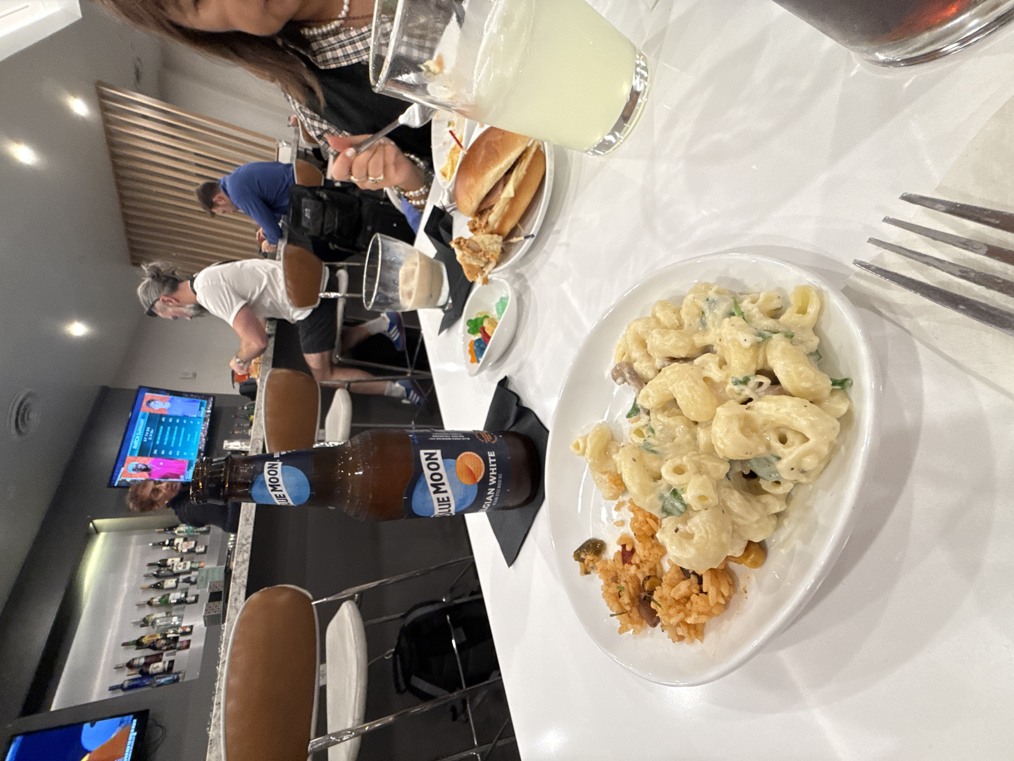 This media file captures an indoor scene, likely a bar, lounge, or restaurant, with several people dining and drinking. The primary focus is on a white table laden with various food and beverage items.
In the foreground, a white plate holds a serving of creamy macaroni and cheese with green flecks, alongside a small portion of reddish-brown rice. A bottle of "BLUE MOON BELGIAN WHITE" wheat ale, with an orange slice depicted on its label, rests on a black napkin beside the plate. A silver fork is visible on the right.
Further up the table, a partial view of a person with dark hair, wearing a plaid shirt and pearl necklace, is seen, holding a fork. Next to them is a glass containing a light yellow beverage, possibly lemonade. Another small plate holds a burger or sandwich and fried food, while a tiny white dish contains colorful gummy candies. A glass with a lighter colored drink is also visible.
In the background, several individuals are present. A man with grey hair tied in a bun and wearing a white t-shirt and grey headband is seen leaning forward. Behind him, a man in a blue shirt is visible. To the left, a bar area is visible with brown-seated stools and shelves stocked with bottles. Two flat-screen televisions are mounted; one displays "ROCK & SUMMER" text and images of people, while the other shows an abstract design. The overall setting appears modern and well-lit by artificial lights. The atmosphere suggests a casual dining or social gathering.