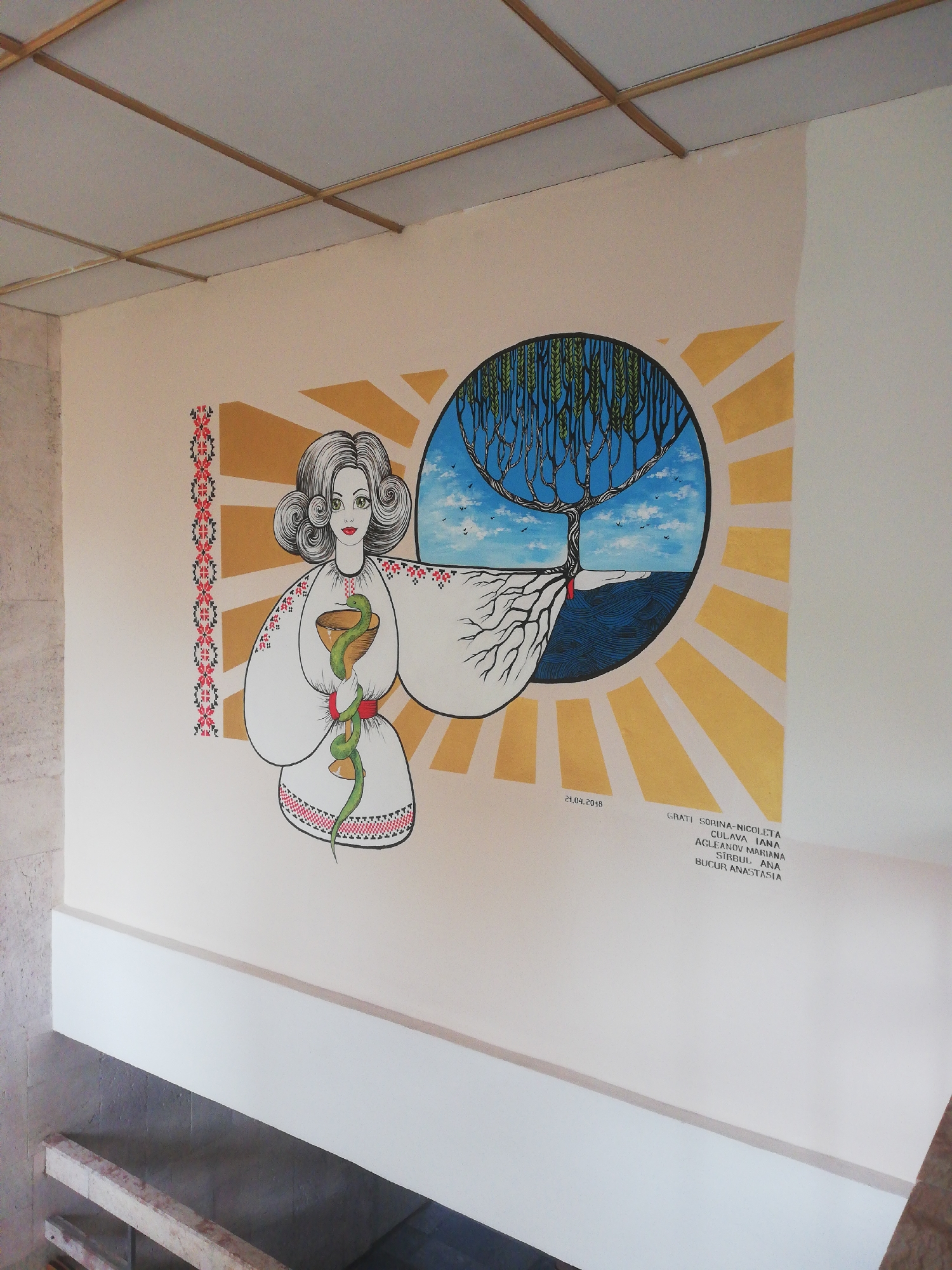 The image shows a mural on a wall, possibly in a public building or institution, located in Bucharest, Romania. The mural depicts a stylized woman wearing a traditional embroidered garment, holding a chalice with a coiled snake around it. Behind her is a circular motif representing a tree with roots extending downwards and branches reaching towards the sky. The sun is represented by radiating golden lines. In the lower right corner of the mural, there is a date "21.04.2018" and a list of names, possibly indicating the artists or individuals involved in the project. The wall is painted a light cream color, and a white ledge runs horizontally across the lower portion of the image. The ceiling above the wall is a suspended grid system with light-colored panels and wooden or metal crossbeams. The overall composition suggests a theme related to health, life, or nature, possibly with cultural or symbolic significance.