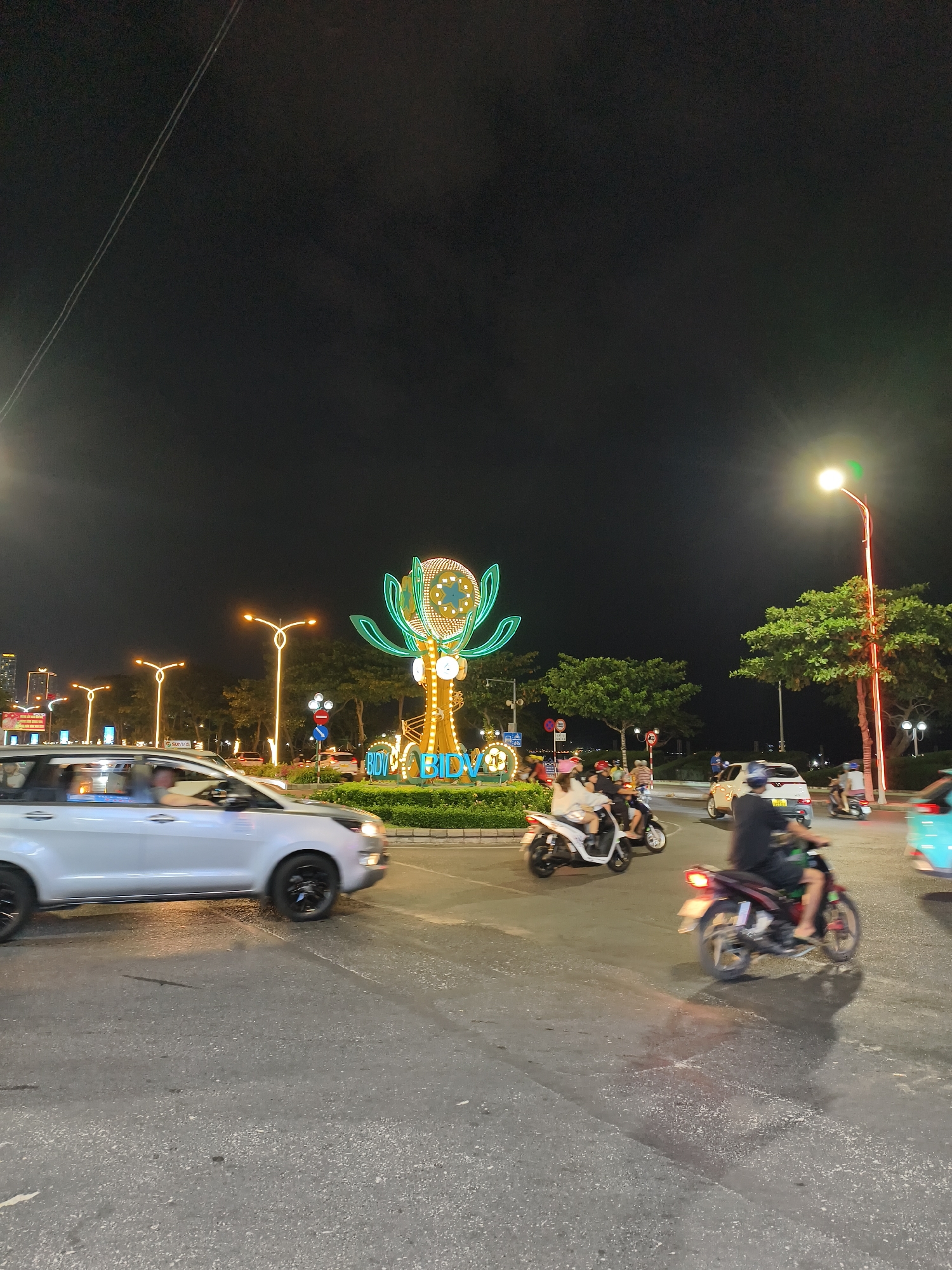 At night, an intersection in Unknown, Unknown shows a busy street with various vehicles. A light-colored car moves to the left of the frame, while several scooters navigate the intersection. In the center of the image, a roundabout features a lighted decorative sculpture. Streetlights illuminate the area, and trees and buildings are visible in the background. The sky is dark, suggesting a clear night.