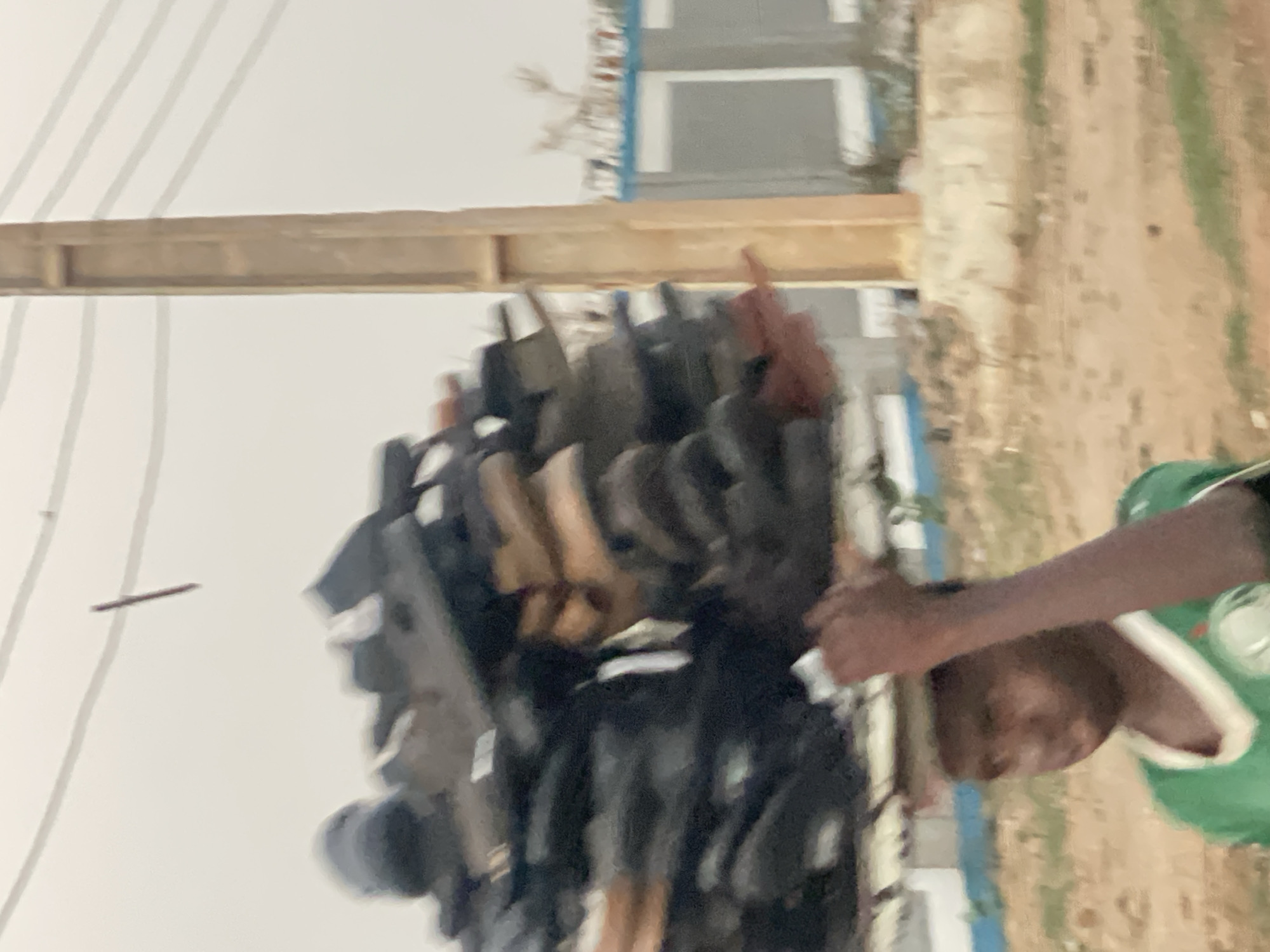 The image depicts a person in a green and white shirt. The background includes a stack of objects, possibly shoes or other items for sale, a wooden beam, a building with a white facade and gray windows, and an unpaved area suggestive of a roadside or market location. The setting is inferred to be Hayin Banki, Nigeria. The person is partially visible. The stack of items appears to be the focus. Electrical wires can also be seen.