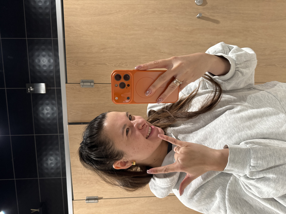 A woman takes a selfie with an orange phone in a public restroom. She is wearing a gray hoodie and has long brown hair. She makes the "rock on" hand gesture with her left hand and smiles. The background consists of black tiled walls with circular patterns and wood paneling with metal hinges and doorknobs. The photo is rotated counterclockwise.