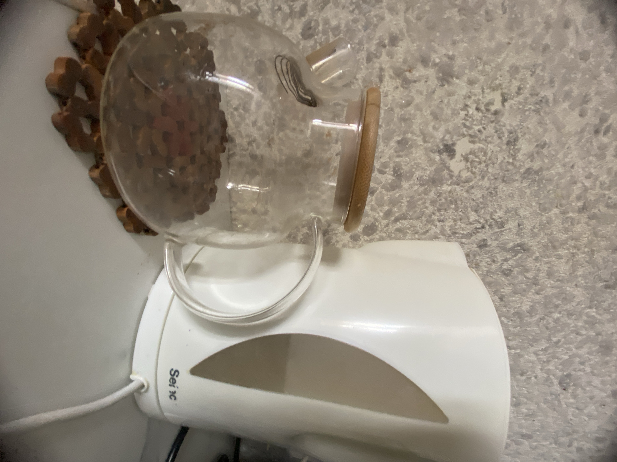 The image depicts a white electric kettle, branded "Sei x", and a glass teapot with a bamboo lid. The teapot is positioned in front of the kettle and contains a dark brown substance, likely coffee beans or tea leaves, resting on a wooden coaster. The background is a textured wall with a mottled gray and white pattern. The objects are placed on a white surface, possibly a countertop or shelf. The scene is indoors, with no specific Ufa landmarks visible.