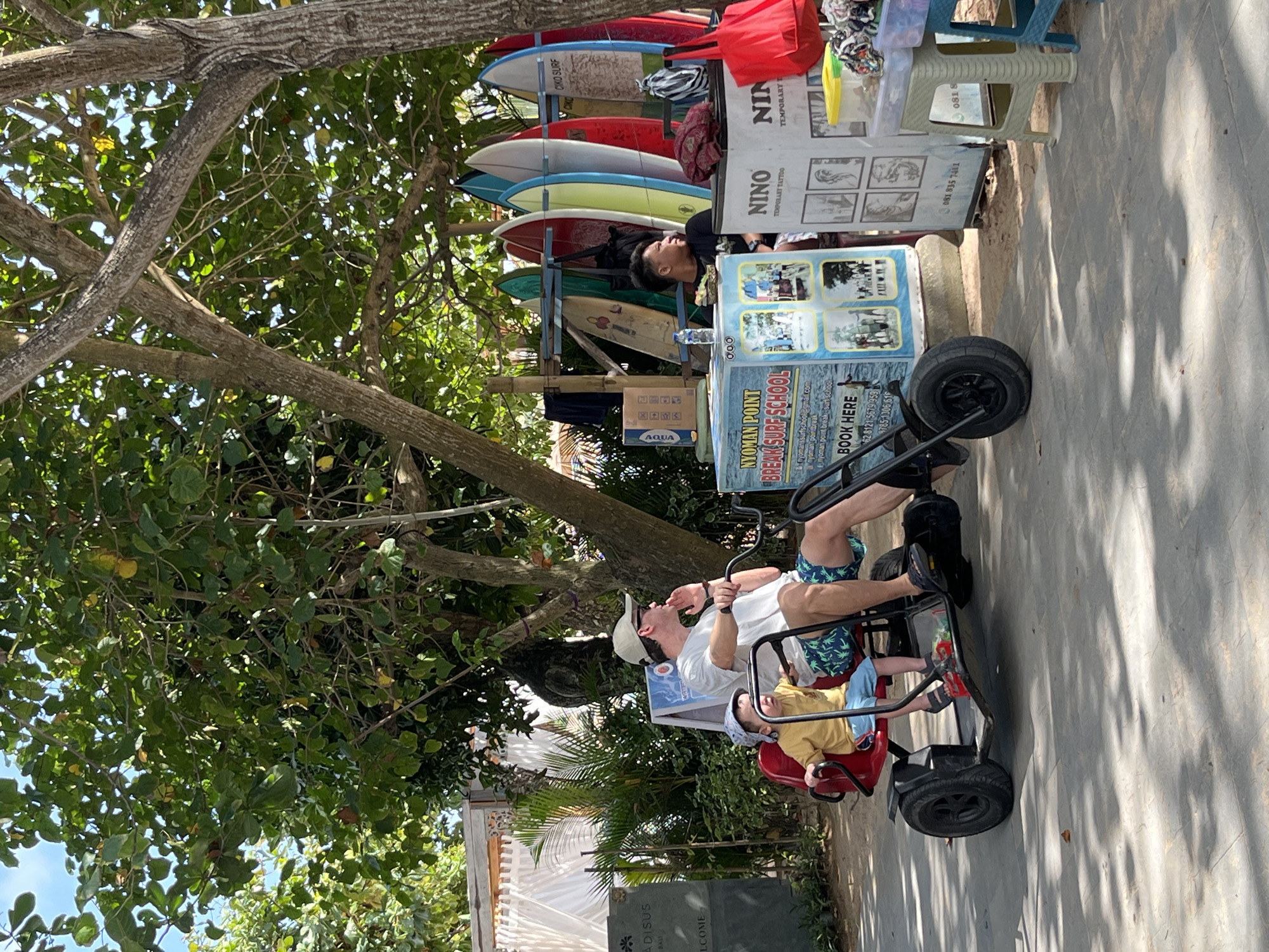 The image depicts a street scene in Kuta Selatan, Indonesia, under a canopy of trees. In the foreground, a man and a child are riding a red pedal-powered go-kart. The man, wearing a white t-shirt, a beige cap, and patterned blue shorts, is steering and pedaling. The child, dressed in a yellow t-shirt and blue shorts, is seated in front of him. To the right, a man in a black t-shirt is sitting and looking to the side. Behind them, surfboards are displayed on a rack, with a stall advertising "NYOMAN POINT BREAK SURF SCHOOL". Further to the right, another stall displays temporary tattoos and various items on shelves. The ground is paved with gray tiles, and shadows from the trees are cast across the scene. In the background, a white tent-like structure and lush green foliage are visible.