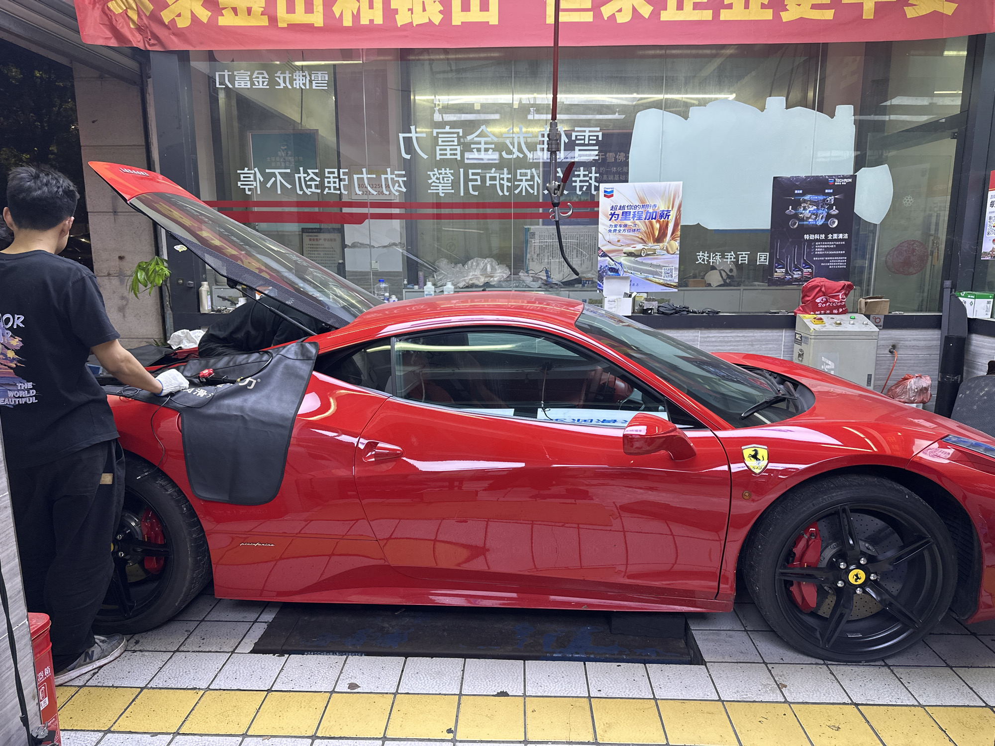 A red Ferrari, likely a 458 model, is parked on a tiled surface, possibly a street-side garage or car detailing shop in Chongqing High-tech Zone, China. The car's rear engine cover is open, revealing mechanical components, and a person is standing next to it, facing away from the camera. The person is wearing a black t-shirt, dark pants, and white gloves on their left hand, appearing to be working on the vehicle's engine. Part of the text on their shirt reads "OR THE WORLD BEAUTIFUL."
The Ferrari features black multi-spoke wheels with bright red brake calipers and yellow Ferrari emblems on the wheels and side panels. The word "pininfarina" is visible on its side bodywork. The vehicle is positioned in front of a glass storefront, reflecting distorted elements from the surroundings.
The shop interior, visible through the glass, contains various items, including bottles, equipment, and posters. A red bag labeled "Softcoo" sits on a counter. A hose with a nozzle hangs down from the ceiling in the reflection.
Visible Chinese text on the shop window includes phrases like "富金山 和银山" (Golden Mountain and Silver Mountain), "永正企业更专业" (Yongzheng Enterprise is More Professional), advertisements for "TECHRON" and "特劲科技 全面清洁" (Techron Technology, Comprehensive Cleaning), and promotions such as "超越你的期待 为里程加薪" (Exceed your expectations, add mileage) and "为爱车做一次 *免费全方位体检" (Give your beloved car a *free comprehensive check-up). A red banner with more Chinese characters hangs above the window. The scene appears to take place during the daytime under diffused light.