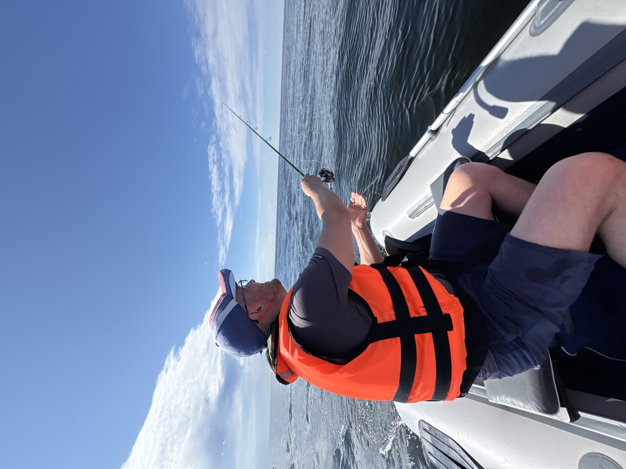 An adult male, wearing a blue and red baseball cap, glasses, a grey t-shirt, and an orange life vest with black stripes, is fishing from an inflatable boat on a body of water. He holds a fishing rod with a reel in his right hand and is looking to his right. The boat, which is grey and white, has black oars visible. The water is dark and appears to be calm with small ripples. The sky above is blue with scattered white clouds. The scene appears to be taking place outdoors, possibly on a lake or the sea. The location is identified as Zelenaya Roshcha, Russia.