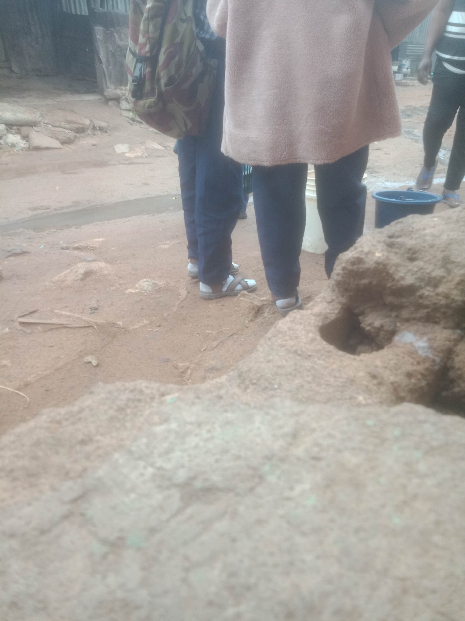 An outdoor medium shot shows the legs and lower torsos of three individuals in a dusty, outdoor environment. The ground is composed of dirt and small rocks, with a shallow puddle of water in the background. One person on the left wears a camouflage backpack and dark blue pants. Another person in the center is wearing a light pink fuzzy jacket over dark blue pants and white sandals. A third person on the right wears black pants with a white stripe detail and appears to be wearing sandals. A dark blue bucket is visible near the center of the frame. In the foreground, a large, rough, tan-colored rock occupies a significant portion of the image. The background shows a weathered structure with vertical corrugated metal siding.