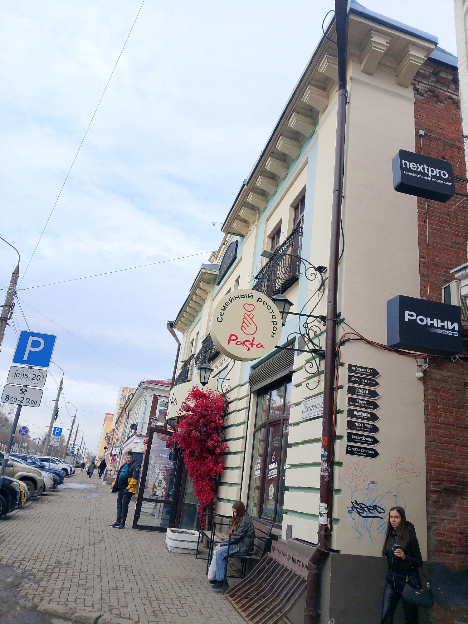 The image depicts a street scene in Izhevsk, Russia. The focus is on a building with a "Pasta" restaurant, indicated by a round sign with a heart-shaped hand gesture and Cyrillic text. Red foliage decorates the storefront.
Two women are visible: one sitting on a bench in front of the building and another standing on the sidewalk holding a cup, possibly coffee. A man stands in the doorway of the restaurant. Cars are parked along the street, marked by a parking sign with time restrictions. Additional signs on the building indicate other nearby establishments such as "Next Pro" (a dance coworking space) and "Ronni Diner." Graffiti is visible on the lower part of the building's wall. The sky is overcast.