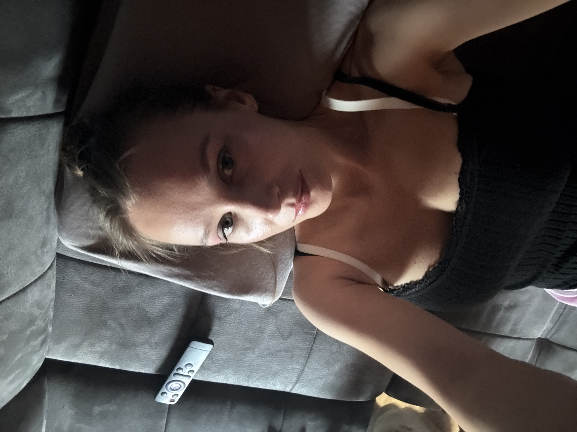 A woman is lying on a grey couch in what appears to be a residential setting in Überherrn, Germany. She is looking up at the camera, with her hair pulled back. She is wearing a black tank top with white bra straps. A silver remote control is visible on the couch to her left. The couch appears to be made of fabric and has a textured surface. There is a pillow behind her head. The lighting suggests it is indoors, possibly at night or in a dimly lit room.