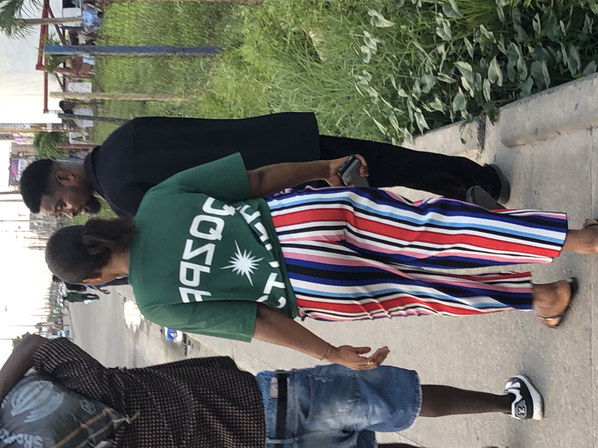 A medium shot of three individuals walking on a concrete sidewalk. One person wears a green t-shirt with white letters and a white starburst design, paired with colorful striped pants and sandals. Another person walks beside them, dressed in a black suit. The third person is wearing a brown and black patterned shirt, blue jean shorts, and black-and-white shoes. In the background, there is greenery and what appears to be a building. The setting is Alaguntan, Nigeria.