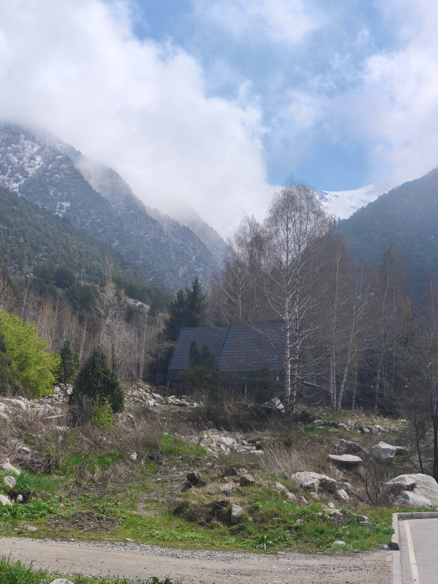 The image depicts a landscape near Kashka Suu, Kyrgyzstan, featuring a small, dark-roofed building nestled among bare birch trees and evergreen conifers. The structure is situated on a rocky, grassy slope interspersed with boulders and dry vegetation. Beyond the trees, the background consists of mountains partially obscured by clouds, with snow visible on the peaks. A dirt road with a concrete edge runs along the foreground. The sky is a mix of blue and white clouds. No people or animals are visible in the scene.