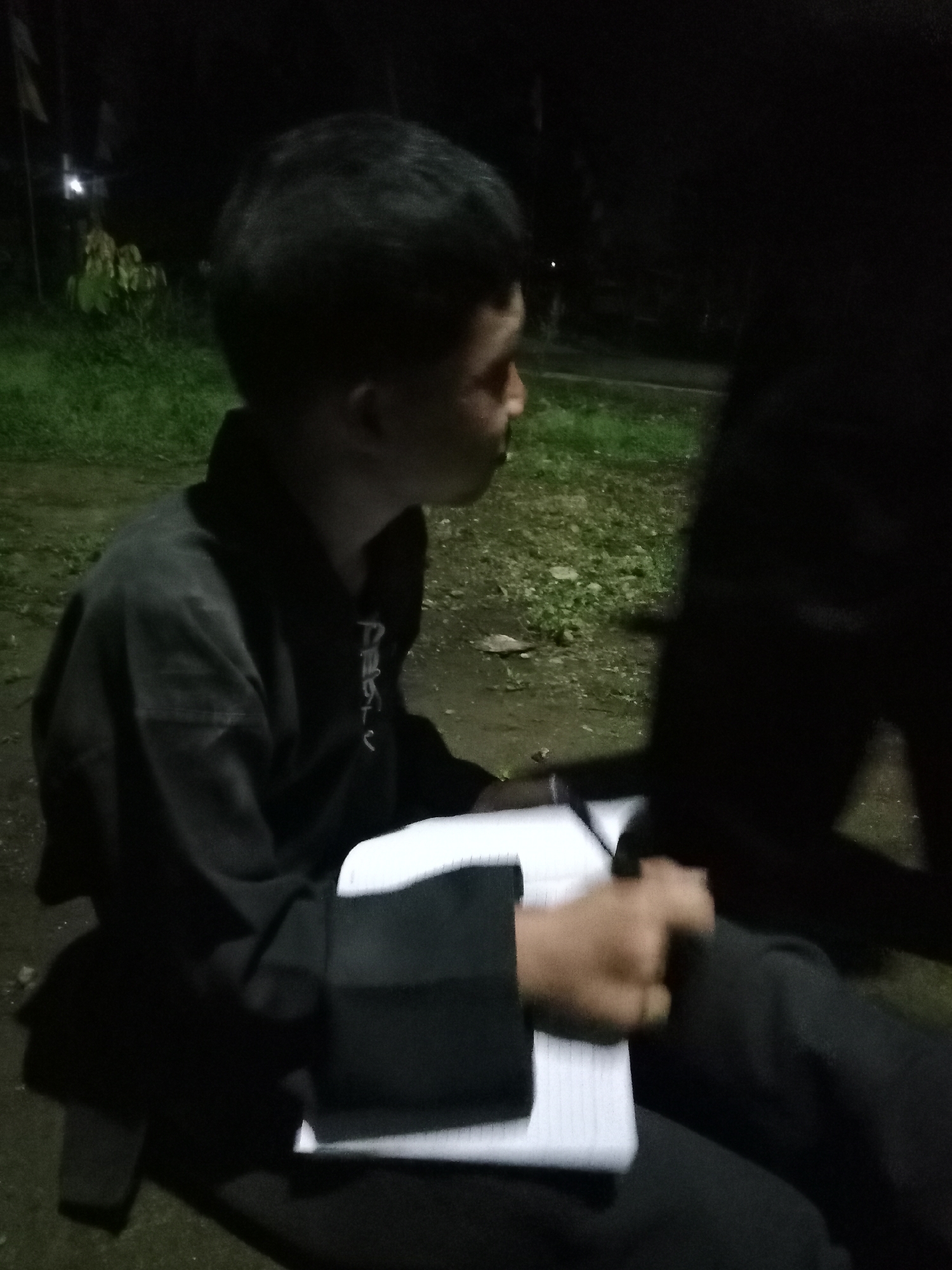 In the city of Liwa, Indonesia, a young person is seated outdoors at night, facing right. They are wearing a black long-sleeved top, possibly a uniform, with some white lettering visible on the chest. The person is holding a white notebook and a pen, suggesting they are studying or taking notes. The background consists of dark foliage and grass, indicating a natural or park-like setting. The lighting is dim, with some ambient light sources contributing to the overall scene. The image captures a moment of quiet activity amidst the nocturnal environment.