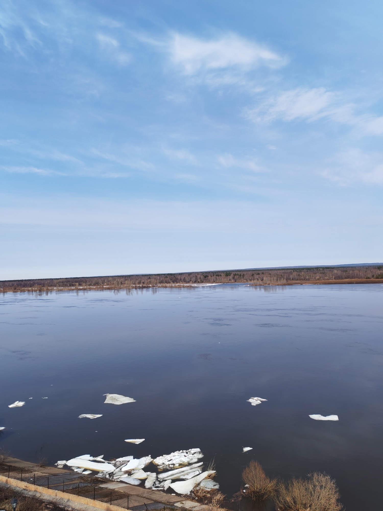 The image depicts a wide river in Sarapul, Russia, under a partly cloudy blue sky. A line of trees is visible on the opposite bank. Ice floes dot the river's surface, with a concentration of larger, broken ice chunks along the near bank. A low wall and fence are visible along the bottom left edge of the frame. Bare bushes and some dry vegetation can also be seen along the near bank. The river appears calm with reflections of the sky.