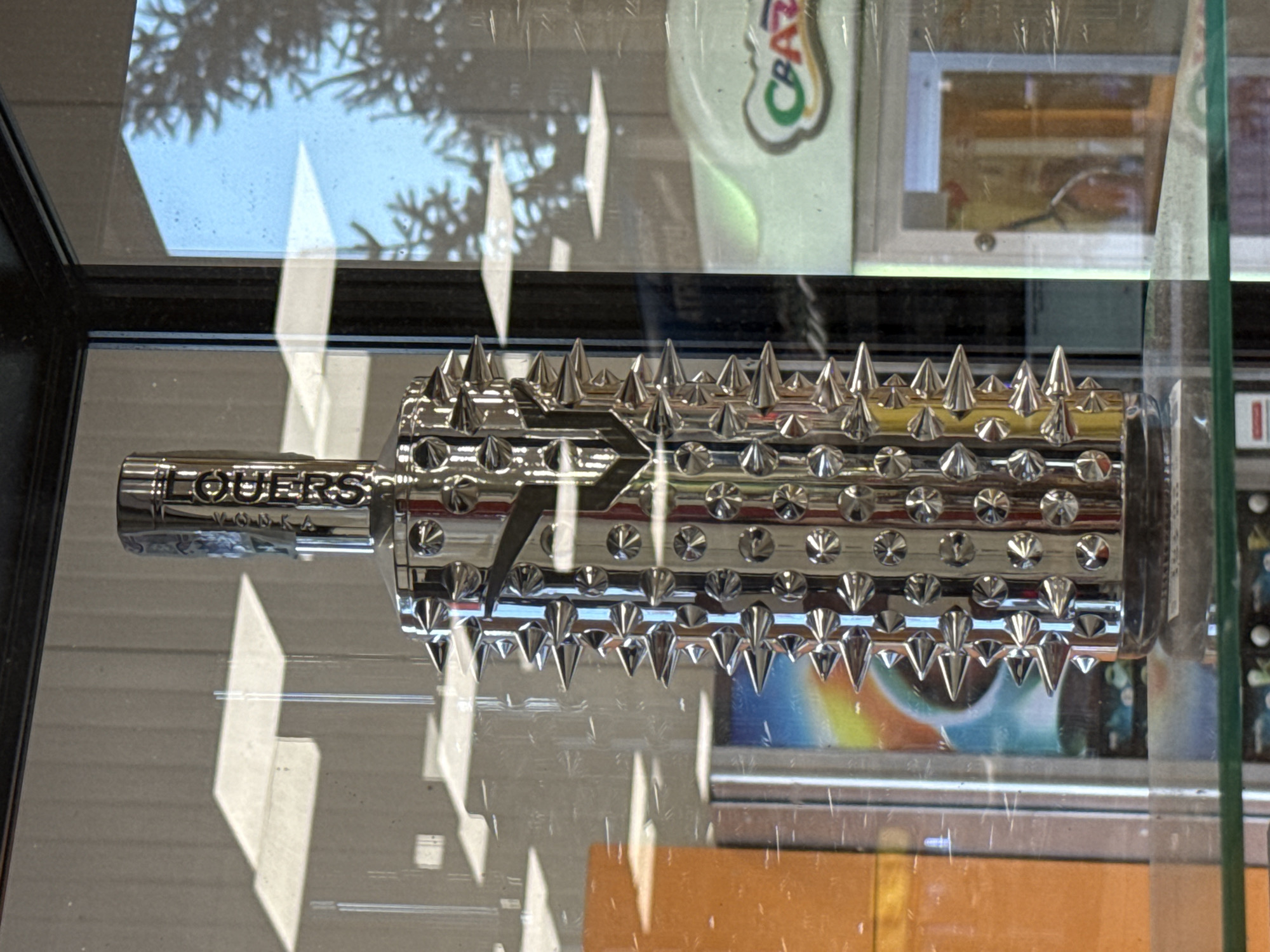 A chrome-plated vodka bottle with sharp, protruding spikes is displayed on a glass shelf inside a display case. The bottle is cylindrical with a faceted cap and is covered in numerous pointed studs, giving it a medieval mace-like appearance. The label on the neck of the bottle is difficult to read due to reflection and a stylized font, but appears to say "VODKA" and possibly a brand name. Behind the bottle, a reflection of a building with vertical paneled siding and large windows is visible, along with the shadow of trees and a bright blue sky. To the right, part of a colorful sign with the word "CRAZY" is visible. The overall environment appears to be a retail store or bar. This image was captured in Montana, Bulgaria.