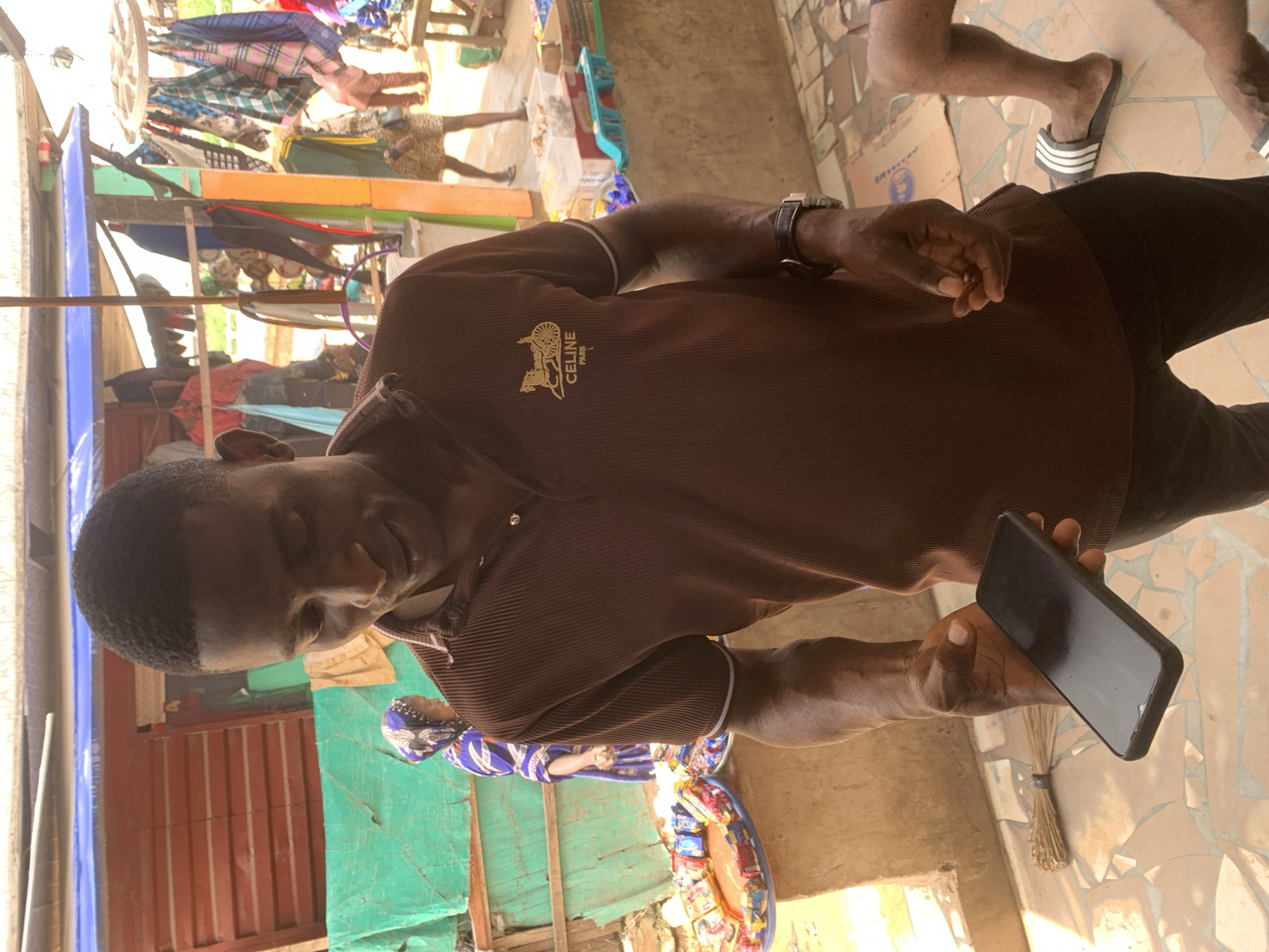 A man in a brown, ribbed polo shirt with a "CELINE PARIS" logo on the left chest, and dark trousers, stands at an outdoor market in Shagamu, Nigeria. He is holding a black smartphone in his left hand. His right hand is extended and appears to be gesturing. He is looking down at the phone. In the background, a woman in a blue and purple headscarf and matching dress is visible behind a display of packaged goods. Other stalls with clothing and various items are also present in the background. The ground is tiled with earth-toned ceramic pieces. A broom is visible leaning against a wall to the left of the man.