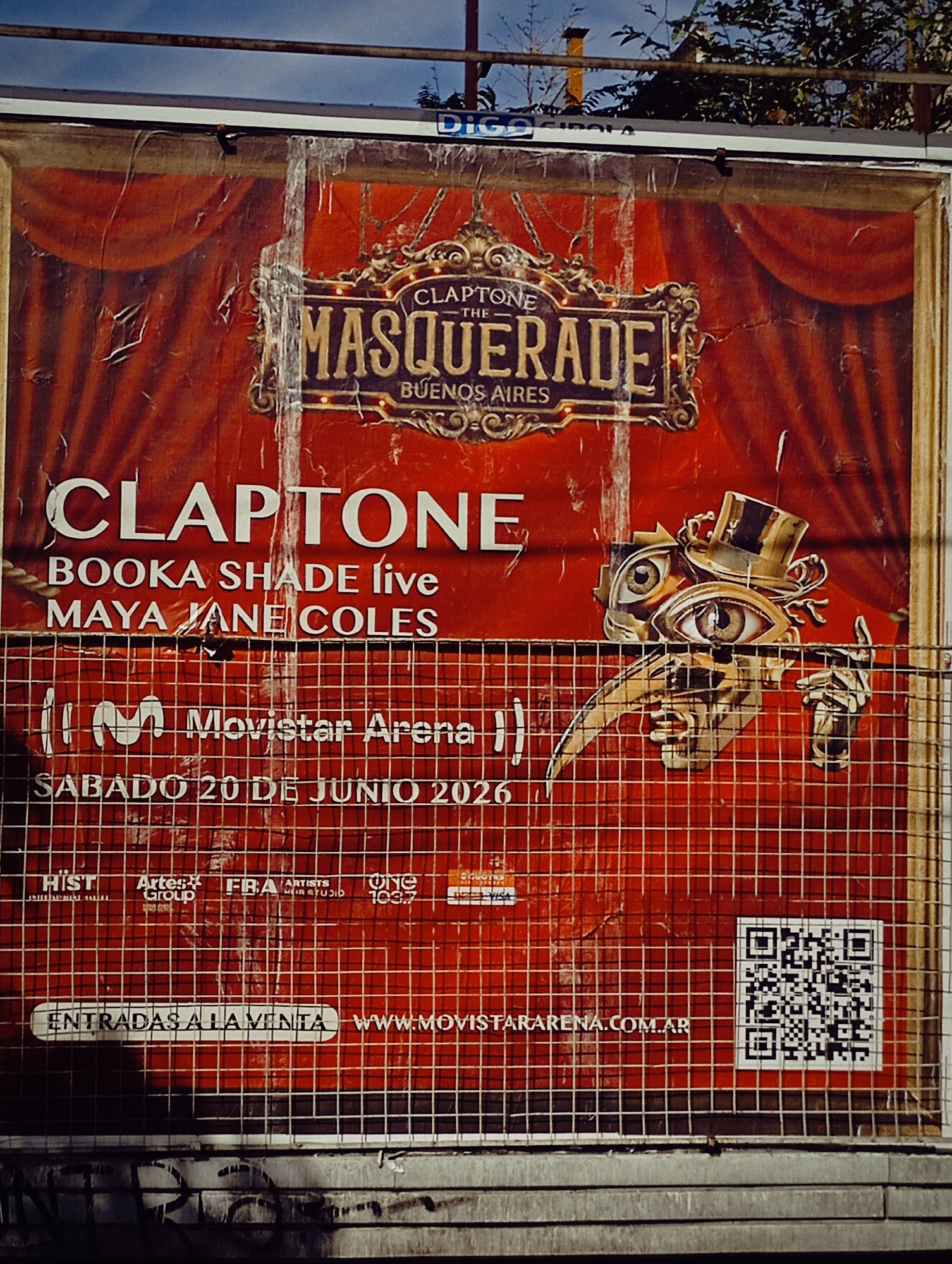 The image shows a weathered advertisement poster in Buenos Aires, Argentina, promoting an event called "Claptone: The Masquerade" at Movistar Arena on Saturday, June 20, 2026. The poster features the names of artists Claptone, Booka Shade (live), and Maya Jane Coles. The poster's design incorporates a mask-like image with eyes, a top hat, and a beak. A QR code is at the bottom right, and the URL "WWW.MOVISTARARENA.COM.AR" is displayed. The poster is affixed to a surface behind a metal grid fence. Various sponsor logos are visible in the lower section of the poster.