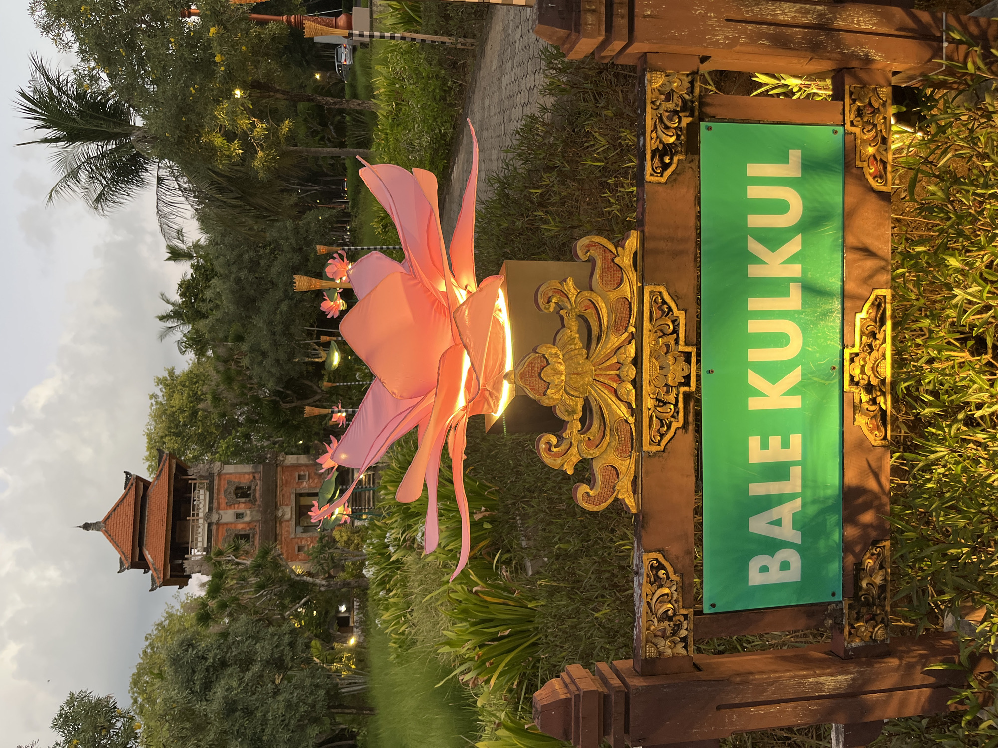 The image depicts an outdoor scene in Kuta Selatan, Indonesia, likely at dusk or dawn, with the sky exhibiting soft, overcast conditions. In the foreground, a decorative wooden sign with ornate carvings frames a bright green rectangular plaque that reads "BALE KULKUL" in white capital letters. This sign is positioned in front of lush green foliage. Behind the sign, a large, illuminated pink lotus-shaped sculpture is prominently displayed, casting a warm glow. Further in the background, a traditional Balinese multi-tiered tower, constructed from reddish brick and wood, stands tall amidst a landscape of trees and manicured greenery. The environment appears to be a public space or park, with a paved pathway visible to the right of the sculpture and trees lining the path. There are no people visible in the frame, and the overall atmosphere is serene and decorative.