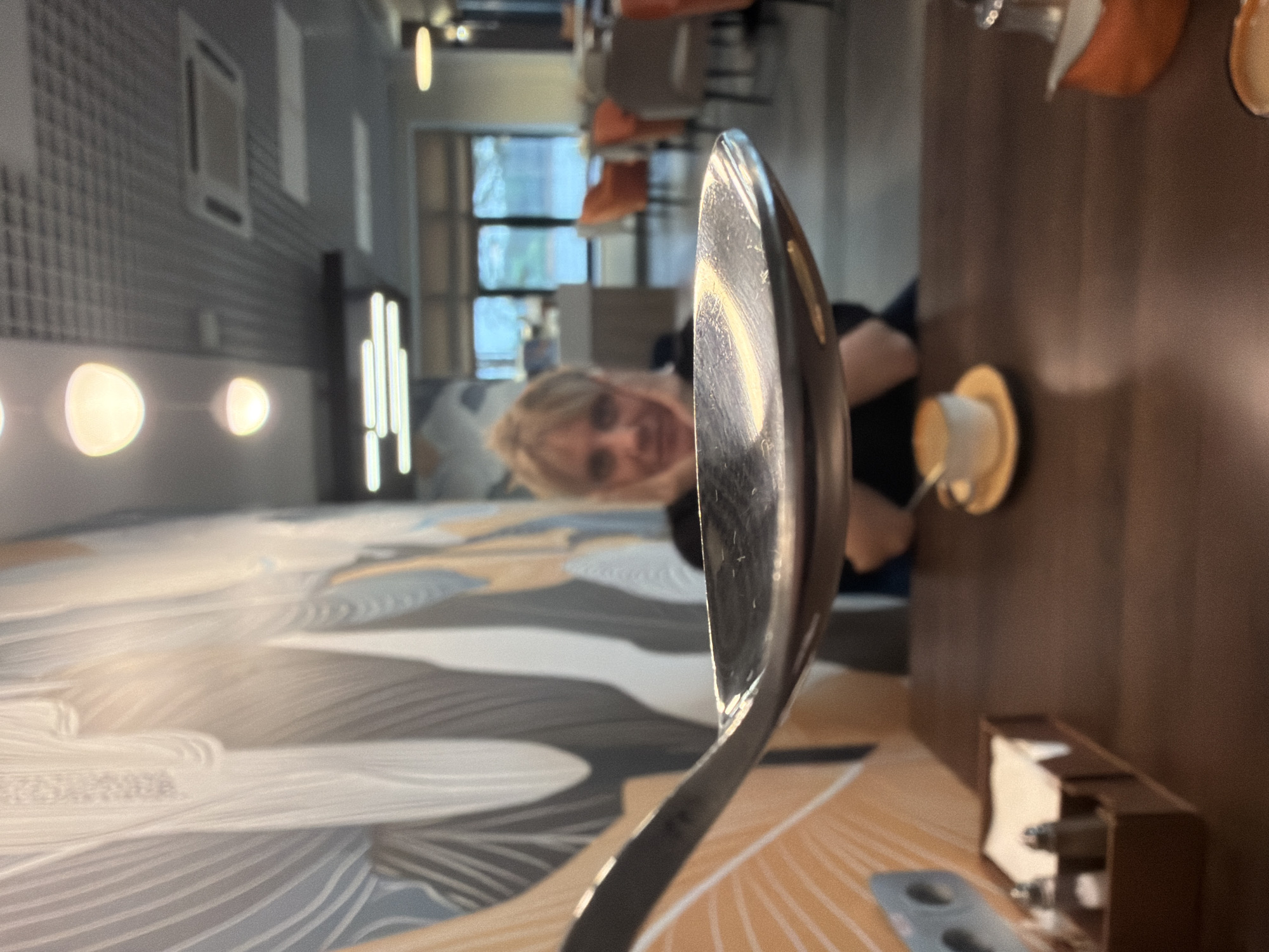 The image, taken in Chaikovsky, Russia, shows a distorted perspective focused on a large, reflective metal spoon or ladle in the foreground. The spoon, positioned vertically on the right side of the frame, reflects parts of the scene.
Reflected and visible directly behind the spoon is a blonde-haired woman with a pleasant smile, looking towards the viewer. She appears to be seated at a table. On the dark brown table to her right, a white cup rests on a saucer. Further to the right and closer to the foreground, a wooden holder containing packets or napkins is visible.
The setting appears to be an indoor dining area, possibly a cafe or restaurant. The background features a wall with a textured, possibly tiled, pattern on the left, adorned with several glowing, oval-shaped lights and vertical strip lights. Natural light streams in from windows further back, reflecting off the furniture. Orange upholstered chairs are partially visible in the background. The floor or a large table surface in the immediate foreground displays an abstract, flowing pattern in shades of grey, white, and light orange-brown. The overall lighting suggests it is daytime. No discernible text is visible. The smiling expression of the woman conveys a relaxed and positive atmosphere.