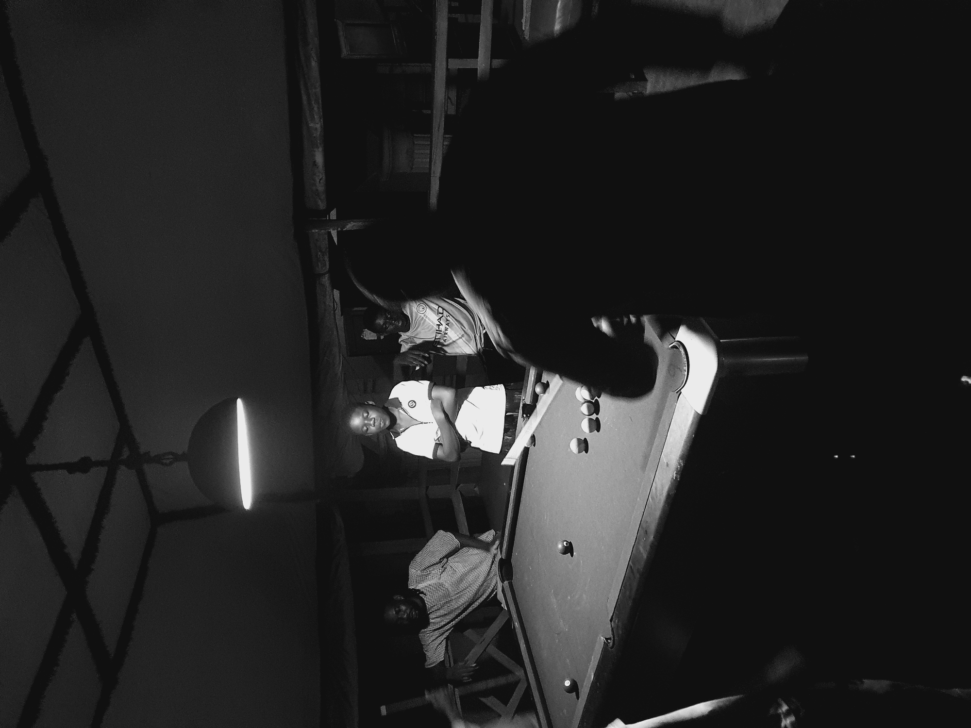 In a dimly lit room, likely an outdoor or semi-outdoor establishment in Lokoja, Nigeria, four individuals are gathered around a pool table. The scene is illuminated by a single overhead lamp, casting long shadows and highlighting the textured ceiling beams. On the left, a man in a striped shirt leans on the pool table, looking towards the right. Facing him, standing with arms crossed, is a young person wearing a white polo shirt with a logo. To their right, another young person wears a light-colored t-shirt with "ETHAD AIRWAYS" visible on it. The fourth individual is a silhouette in the foreground, their back to the camera, suggesting they are either playing or observing the game. Several billiard balls are scattered across the green felt surface of the pool table. The overall atmosphere suggests a casual gathering or a local hangout spot.