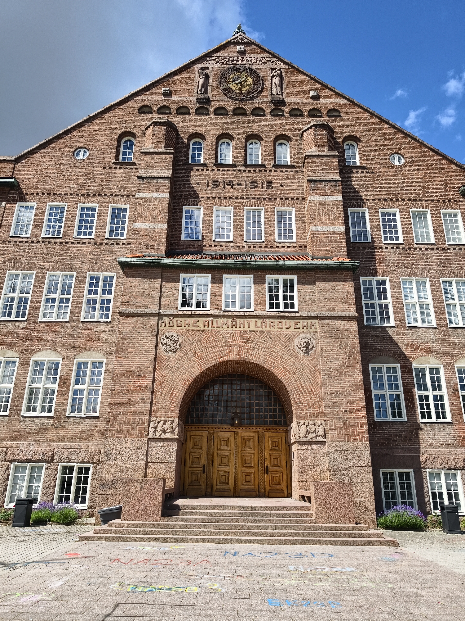 The image shows the main entrance of the "Högare Allmänt Läroverk" in Linköping, Sweden. The building is constructed of red brick with multiple floors and many windows. The entrance features a large arched doorway with double wooden doors, accessed by a wide stone staircase. Above the entrance, inscribed text reads "HÖGRE ALLMÄNT LÄROVERK". The building's facade is adorned with architectural details, including decorative carvings and a prominent clock face near the apex of the roof. In front of the building, the ground is paved with stone tiles, and there are chalk drawings on the pavement. There are no people visible in the image. The sky above is partly cloudy.