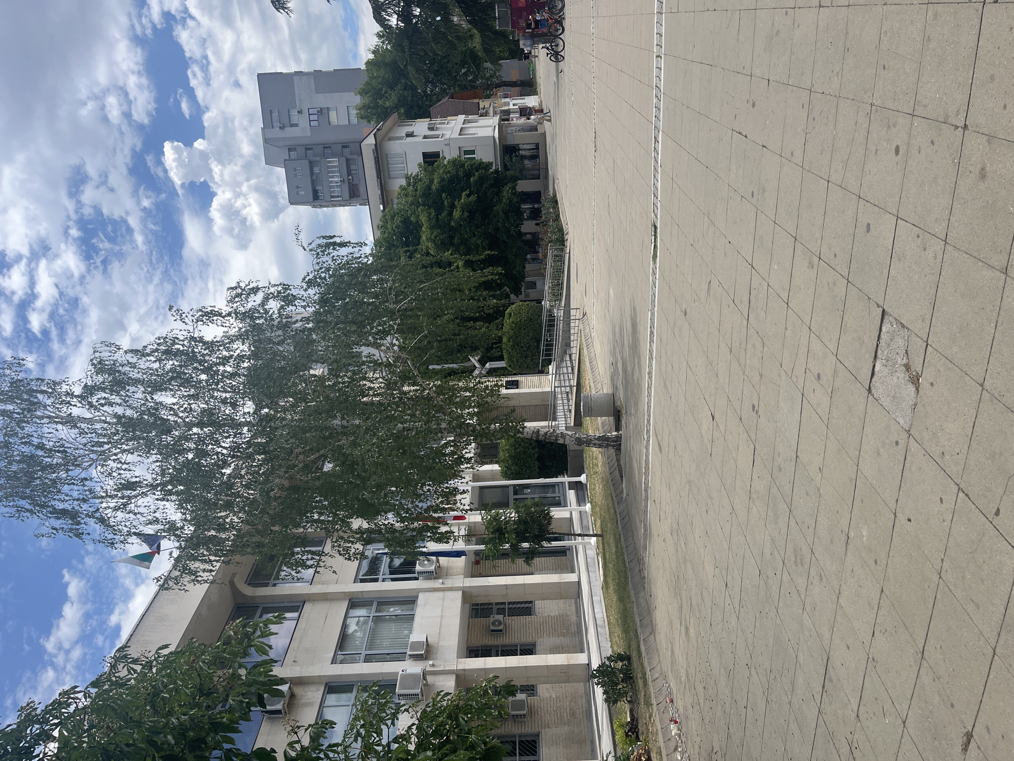 The image depicts a view in Blagoevgrad, Bulgaria. The foreground is dominated by a wide pedestrian walkway paved with grey concrete slabs. A large tree with green foliage is on the left side of the frame, partially obscuring a multi-story building with a light-colored facade and multiple windows, some with air conditioning units. The flag of Bulgaria is visible atop the building. Further back, a mid-rise grey building can be seen. In the background, other buildings and trees are visible along with a few bicycles. The sky is blue with scattered white clouds.