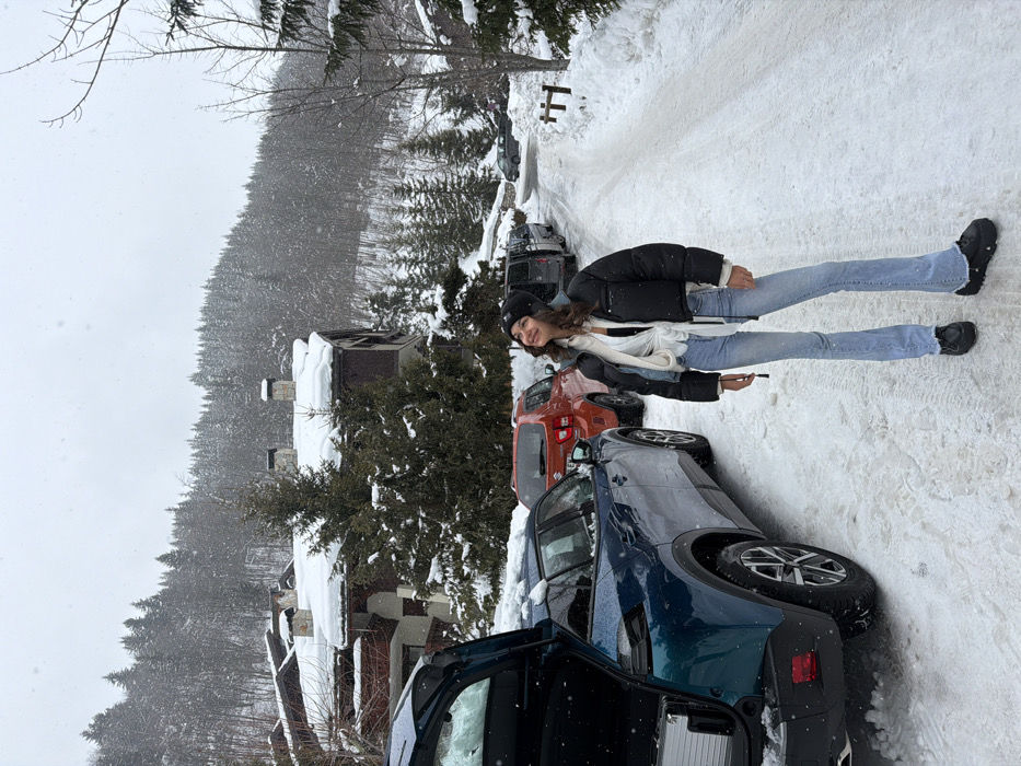 The image depicts a snow-covered scene in Les Allues, France, under an overcast sky. A young adult female stands on a snow-covered road, looking upwards with a slight smile. She wears a black beanie, a black cropped puffer jacket with white lining, a white tied shirt, light blue flared jeans, and black chunky boots. To her left, a dark teal SUV with an open rear tailgate is parked on the snowy road, revealing a silver rectangular object inside its cargo area. Behind this vehicle, an orange Suzuki Ignis and other dark vehicles are parked. In the background, multiple chalet-style buildings with snow-covered roofs and chimneys are visible, nestled amidst dense, snow-laden evergreen and deciduous trees that cover a sloping hillside. Snowflakes are discernible in the air. The ground is uniformly covered in fresh snow, with tire tracks visible on the road.