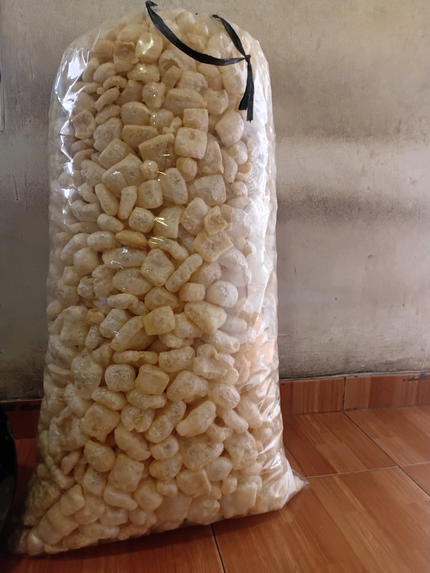 A large, transparent plastic bag, tied at the top with a black ribbon, is filled with a light-colored, irregularly shaped, puffed snack food. The bag is standing upright, leaning slightly against a textured, light-colored wall. The floor beneath the bag is tiled with light brown, rectangular tiles. The scene appears to be indoors, possibly in a storage area or market stall, in the city of Tangerang, Indonesia.