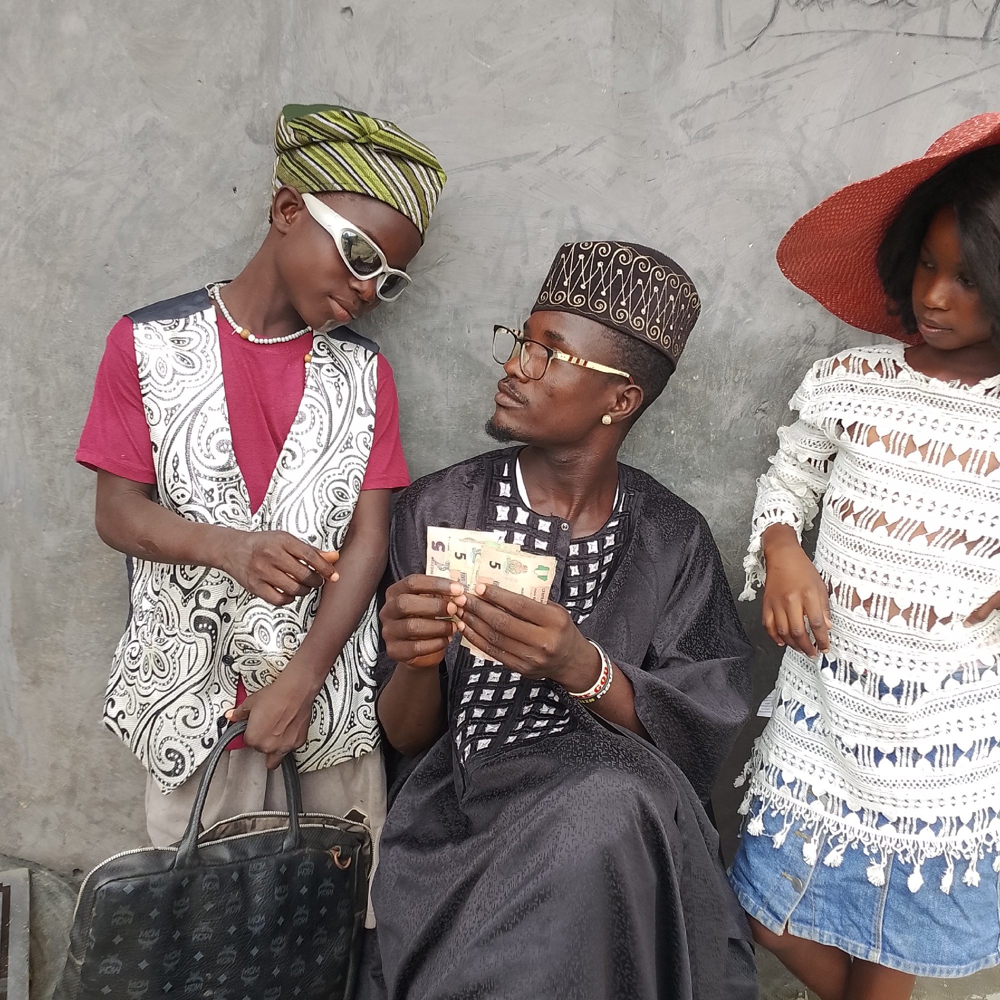 In Ado, Nigeria, three individuals are positioned against a textured gray concrete wall. On the left, a young person wearing a green and white striped cap, white sunglasses, and a patterned vest over a maroon shirt leans against the wall. A beaded necklace is visible. In the center, an adult wearing a black traditional outfit with patterned accents, a black cap with gold embellishments, and glasses holds several Nigerian Naira banknotes in their hands. They are looking towards the young person. On the right, a young person wearing a wide-brimmed red hat and a white crocheted top with a denim skirt stands looking down. A black MCM-patterned bag rests on the ground in front of the young person on the left. The lighting suggests an outdoor, daytime setting.