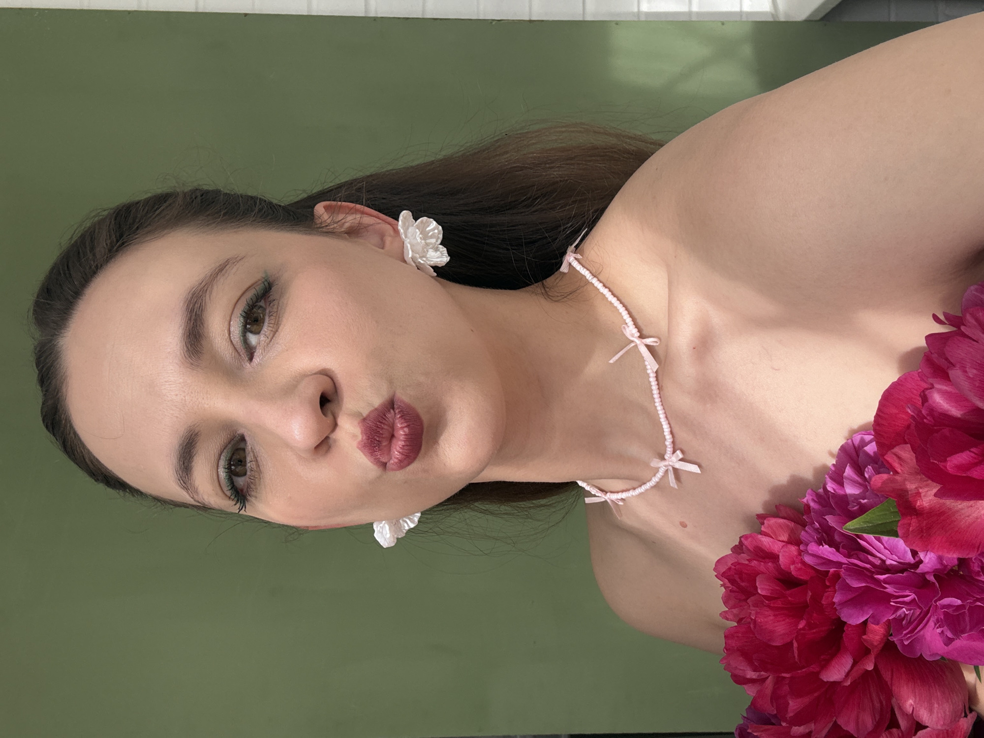 A woman with brown hair pulled back, wearing white flower-shaped earrings and a pink beaded necklace with bows, puckers her lips. She has green eyeliner and dark pink lipstick. She is holding a bouquet of bright pink and magenta peonies in front of her bare shoulders. The background is a solid, muted green wall. The lighting suggests an outdoor setting, possibly on a sunny day. The image does not provide any specific information about the location in Chaikovsky, Russia.