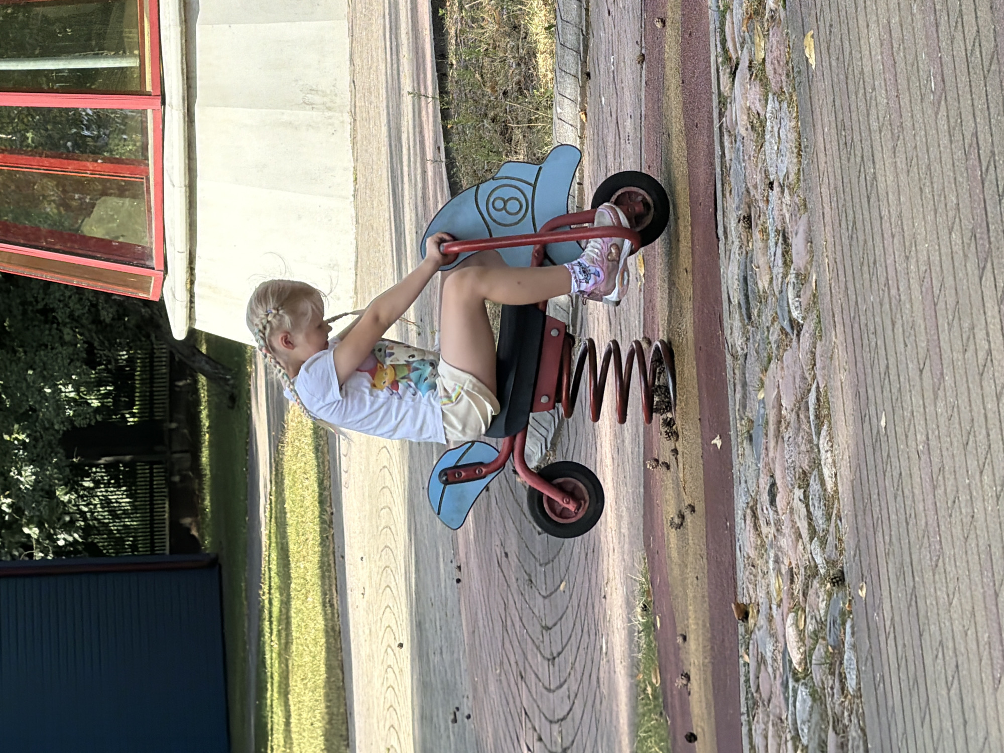 A young girl with blonde braided hair sits on a red and blue spring rider toy shaped like a motorcycle in a park in Ventspils, Latvia. She is wearing a white t-shirt with a colorful cartoon print and light-colored shorts. The girl is looking down, with her hands resting on the handlebars of the toy. The spring rider is positioned on a paved surface made of interlocking bricks with a section of cobblestones visible nearby. The background features green grass, trees, and a portion of a modern building with red trim. Fallen pine cones are scattered on the ground around the toy and on the paved surfaces. The scene appears to be a sunny day.