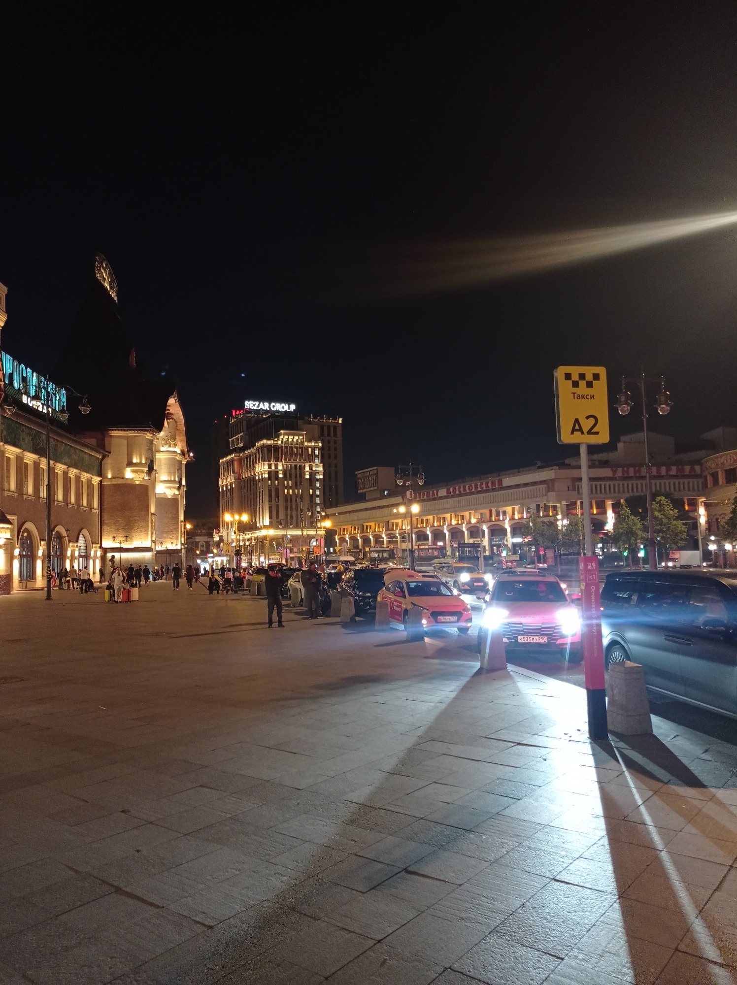 The image depicts a street scene at night in Moscow, Russia, likely near a transportation hub given the presence of taxis and a large building with illuminated signage. On the left, a long, multi-story building with arched windows and illuminated sections is visible, resembling a train station or a historical building. The signage above it appears to be in Cyrillic. In the mid-ground, a modern, well-lit building with the prominent signage "SEZAR GROUP" stands tall. The street is paved with large square tiles and is illuminated by streetlights and the lights from the buildings and vehicles.
Several cars are parked and in motion on the right side of the street. A yellow taxi sign with "Taksi" and "A2" is prominently displayed on a pole in the foreground. Among the vehicles, there are several taxis, including a pink one with Cyrillic license plates and a red taxi with a visible license plate. People are seen walking on the sidewalk in front of the buildings, some with luggage, suggesting travel. The night sky is dark, with a bright light beam or lens flare cutting across the upper right portion of the image. The overall atmosphere is urban and bustling despite the late hour.
