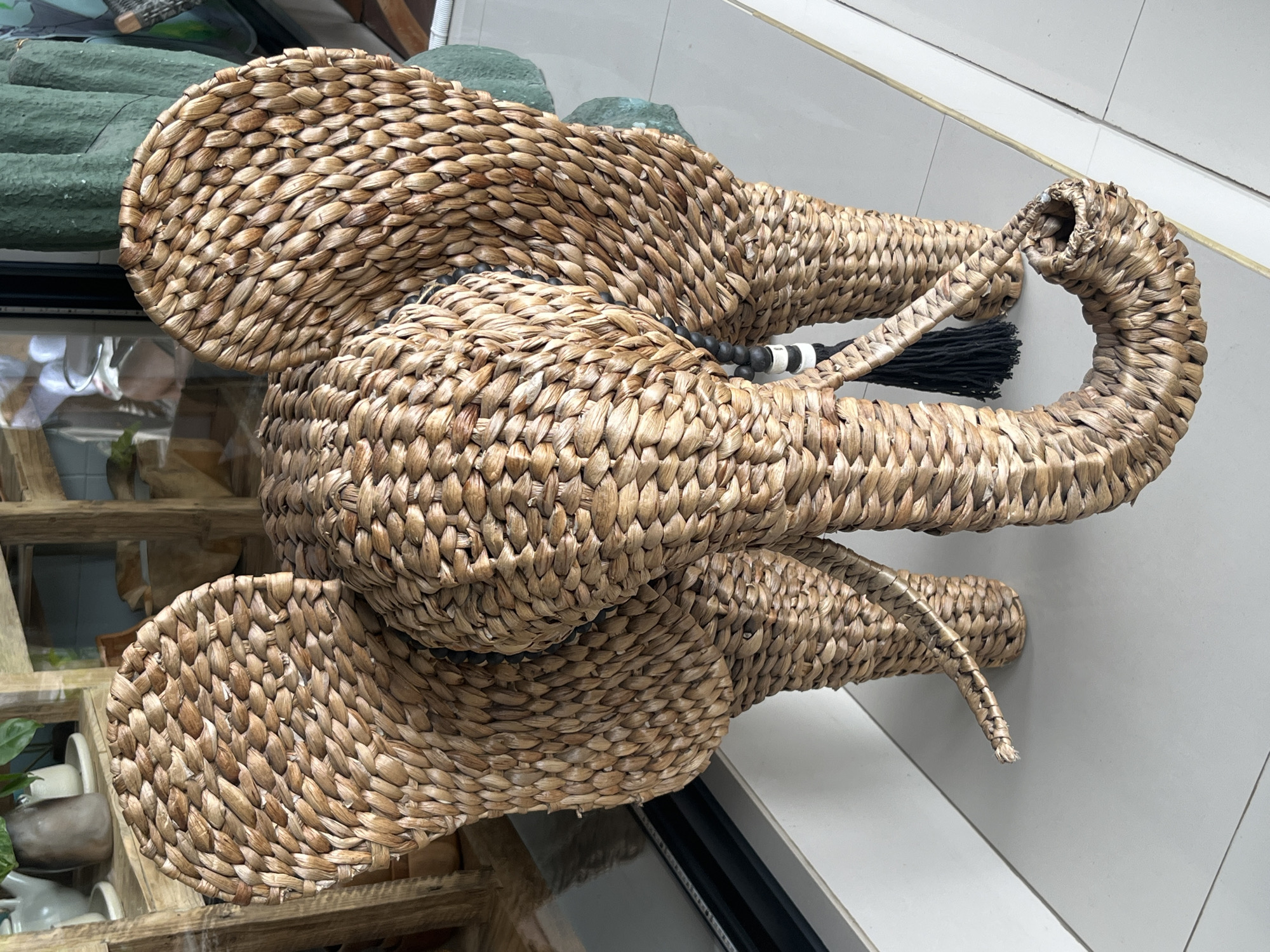 A woven rattan elephant figurine stands on a white tiled floor. The figurine is made of natural, light brown rattan material and is intricately woven to represent an elephant with large ears, a trunk, tusks, and four legs. A black tassel hangs from the side of the elephant, and black and white beads adorn its neck. In the background, a wooden shelf unit with various decorative items is visible, along with a large green textured object that appears to be a decorative plant or sculpture. The scene is indoors, likely in a shop or home, given the display of decorative items. The location is Kuta Selatan, Indonesia.