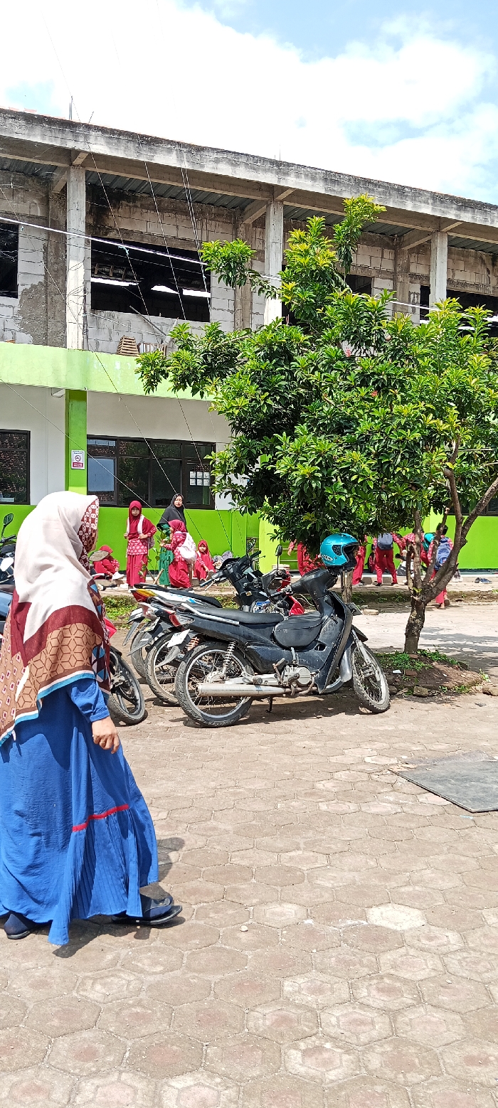 The scene is in front of a concrete building with green painted walls and windows. It is located at coordinates -7.065 latitude, 107.552 longitude in Cangkuang, Indonesia. There is a large tree on the right side of the image and a group of people wearing red and black clothing near the building. A woman in a blue dress and head scarf is walking past a group of motorcycles parked in front of the building. The ground is paved with hexagonal concrete blocks. The sky is clear blue with a few white clouds. There is a small sign on the wall of the building. There are no cars or other vehicles visible.