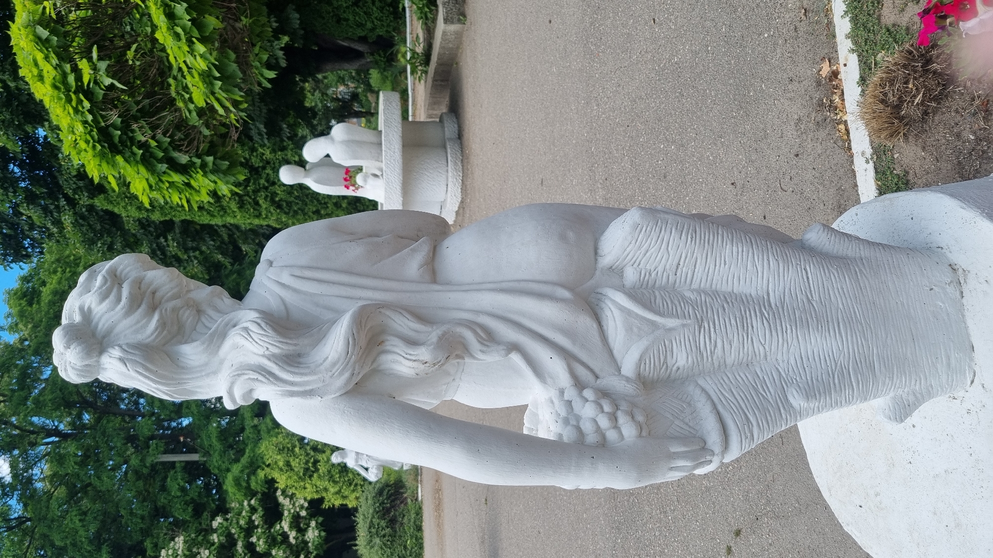 The image displays a white statue of a woman from behind, seen in a park setting. The statue depicts a woman with her hair styled in an updo and long, flowing curls. She is draped in fabric, and her left arm is bent, holding a bunch of grapes. The statue appears to be made of concrete and is mounted on a white pedestal. In the background, there are trees, other statues, and a paved walkway. Further in the background, another white sculpture of two figures leaning towards each other can be seen. The overall scene is of an outdoor public space, likely a garden or park. The context of Smila, Ukraine is not visually apparent from the image itself.